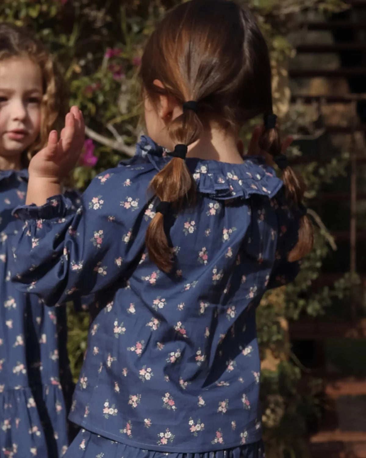 Early Sunday, Cécile Blouse in Ditsy Bouquet Navy, made from Printed Cotton Satin fabric, featuring a relaxed silhouette with gathered sleeves and delicate detailing, shown in a back view of model playing outdoors
