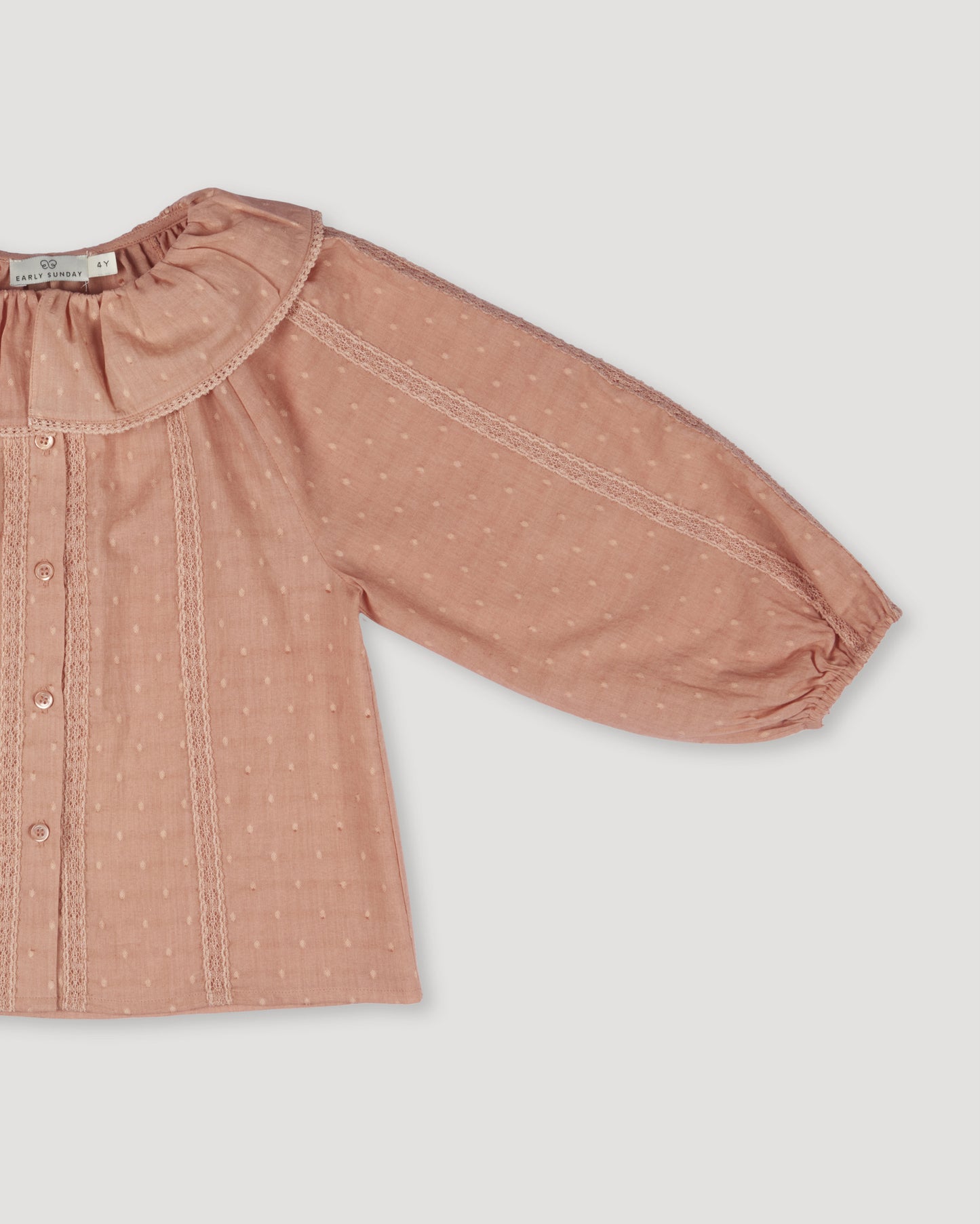 Early Sunday, Cecilia Blouse in Earthy Pink color, made from Cotton Swiss Dobby fabric, featuring relaxed fit, frill neckline, long sleeves with elasticated cuffs, and MOP buttons, shown in a closeup flat-lay view