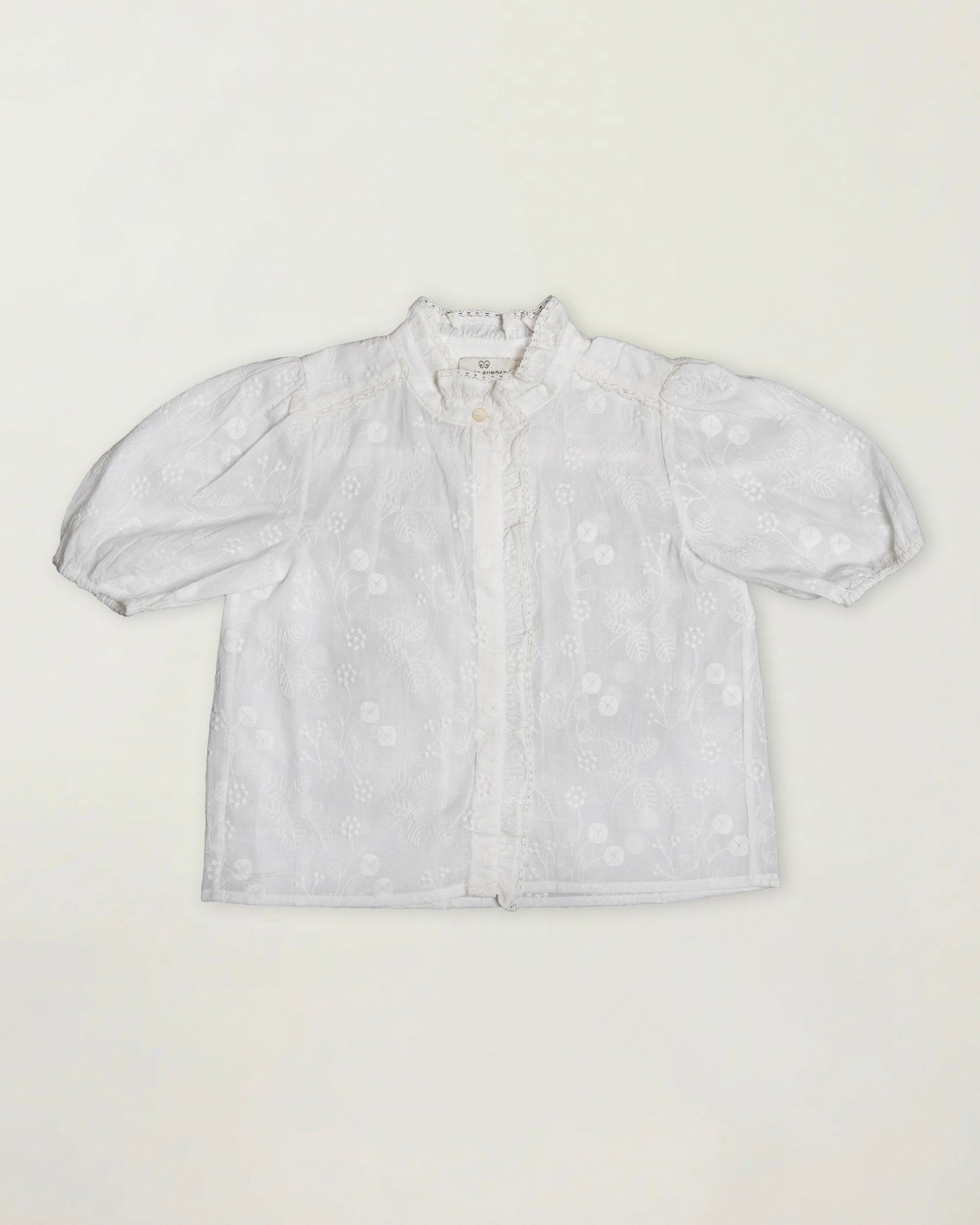 Early Sunday, Coco Blouse in Ivory color, made from all over embroidery fabric, featuring Puff sleeves, ruffled neckline and placket, shown in a front flat-lay view