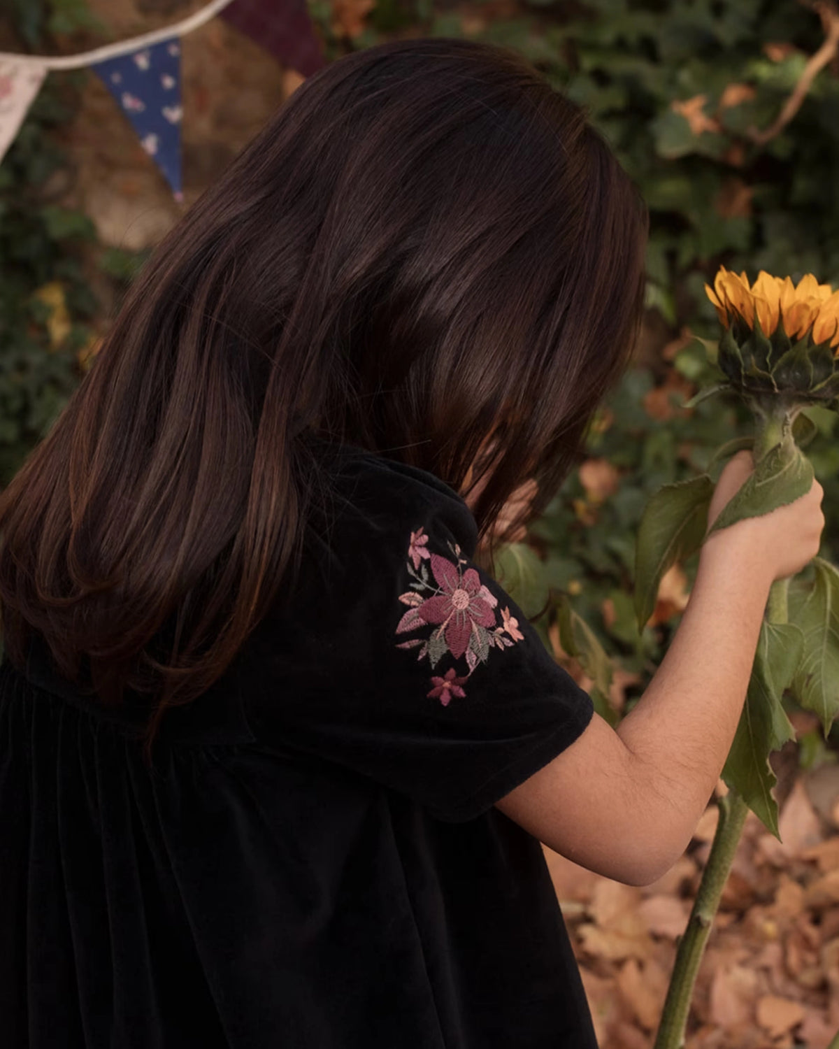 Early Sunday, Erika Dress in Black, made from Cotton Velvet Velour's fabric, featuring a beautiful embroidery with frilled neckline, shown in a side view of model playing outdoors