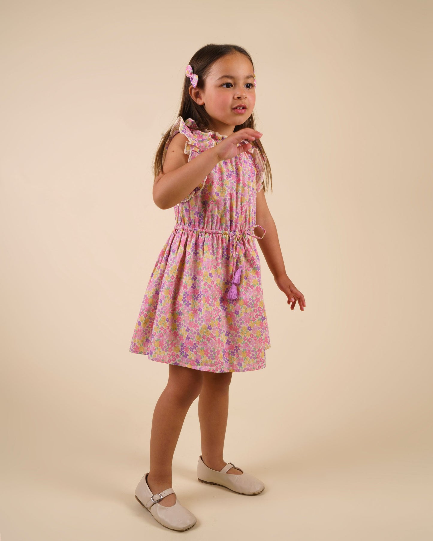 Early Sunday, Florence Dress in Emma Floral Purple color, made from printed cotton cambric fabric, featuring Ruffle detailing on the yoke and sleeves, lace accents, gathered waist with tassel tie, shown in a front view of model wearing