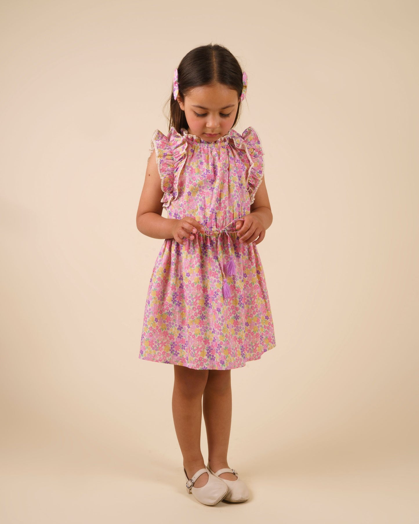 Early Sunday, Florence Dress in Emma Floral Purple color, made from printed cotton cambric fabric, featuring Ruffle detailing on the yoke and sleeves, lace accents, gathered waist with tassel tie, shown in a model standing facing front wearing