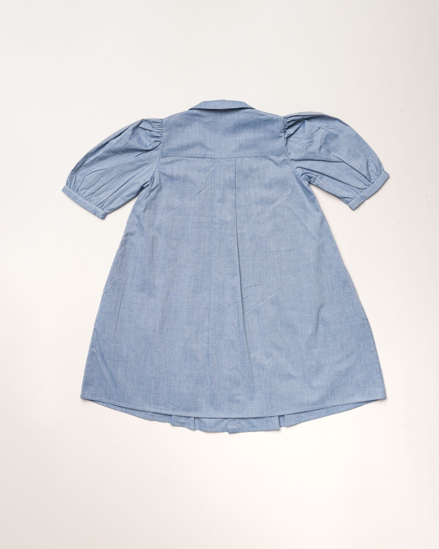 Early Sunday, Jane Dress in Summer Sky color, made from 100% cotton chambray fabric, featuring Smocked / embroidered yoke, peter pan collar and puffed short sleeves, shown in a back flat-lay view