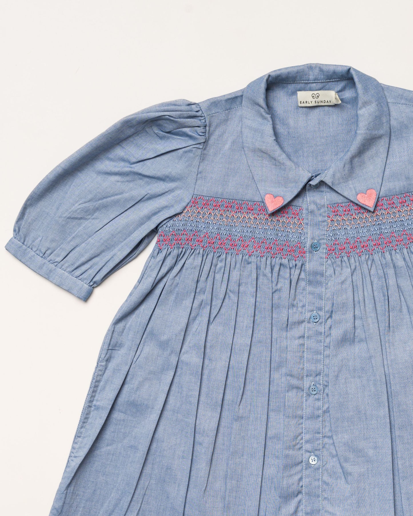 Early Sunday, Jane Dress in Summer Sky color, made from 100% cotton chambray fabric, featuring Smocked / embroidered yoke, peter pan collar and puffed short sleeves, shown in a closeup flat-lay view