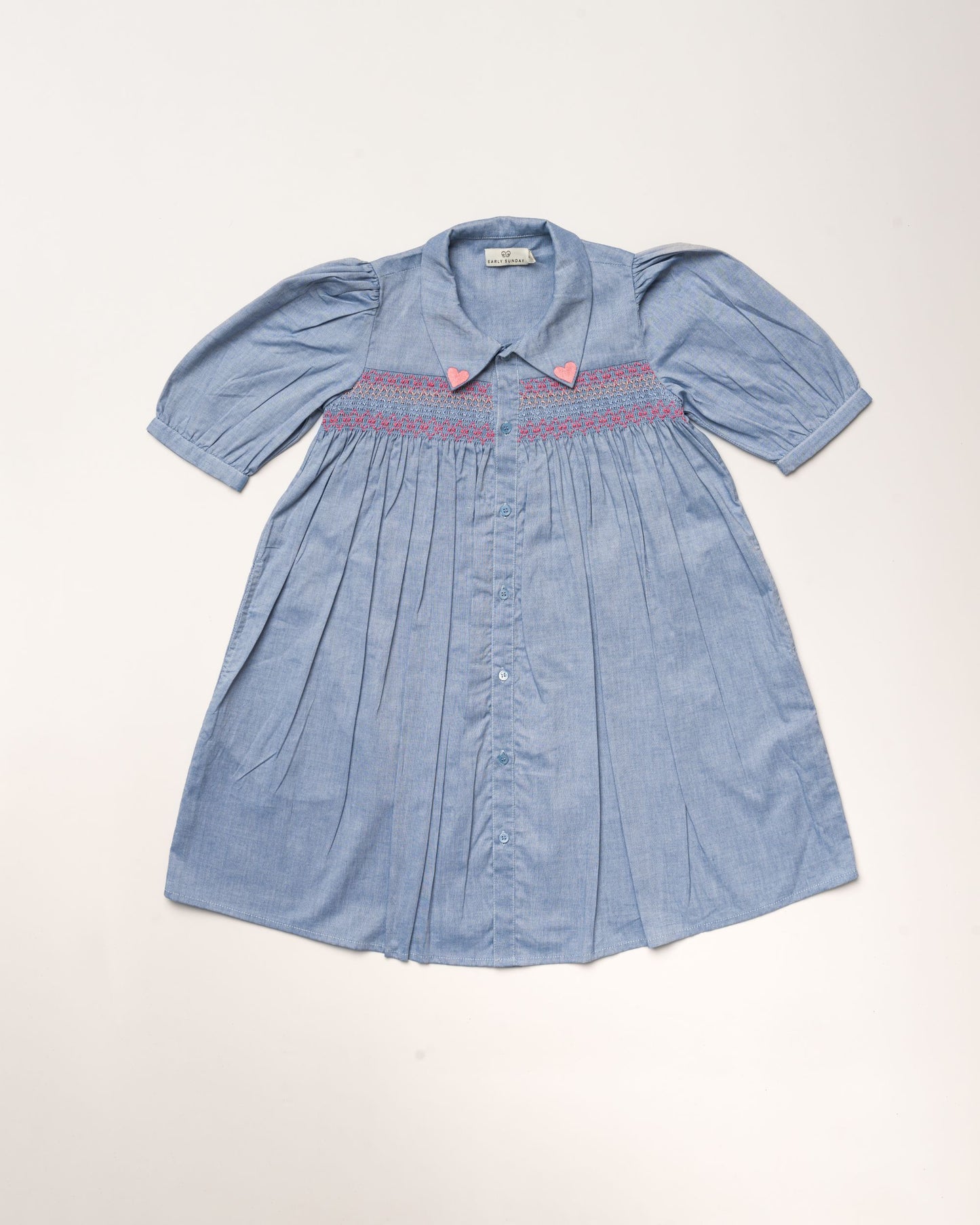 Early Sunday, Jane Dress in Summer Sky color, made from 100% cotton chambray fabric, featuring Smocked / embroidered yoke, peter pan collar and puffed short sleeves, shown in a front flat-lay view