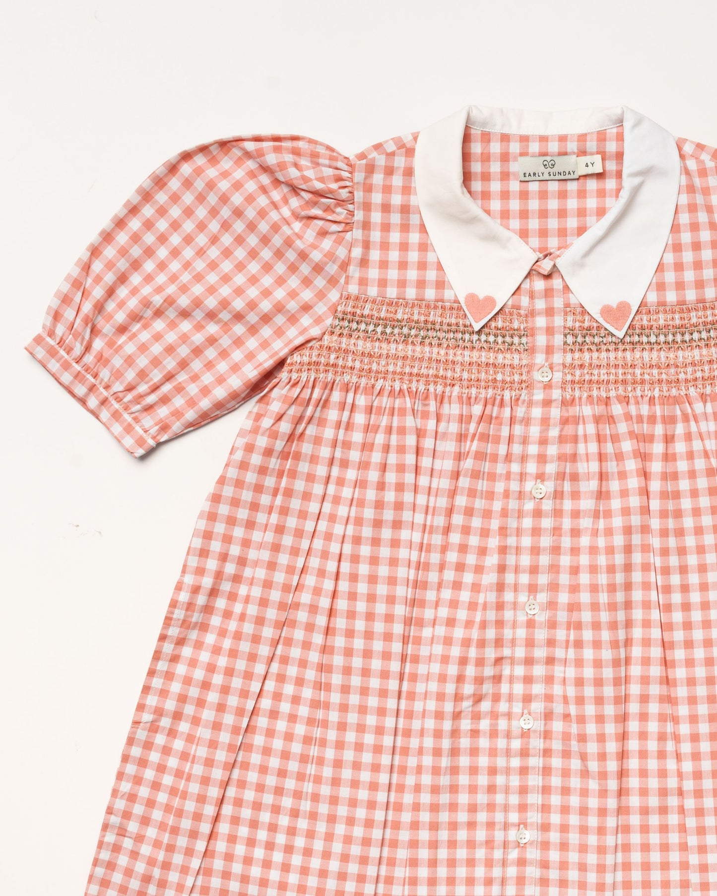 Early Sunday, Jane Dress in Peony color, made from 100% cotton gingham checks fabric, featuring Smocked / embroidered yoke, peter pan collar and puffed short sleeves, shown in a closeup flat-lay view