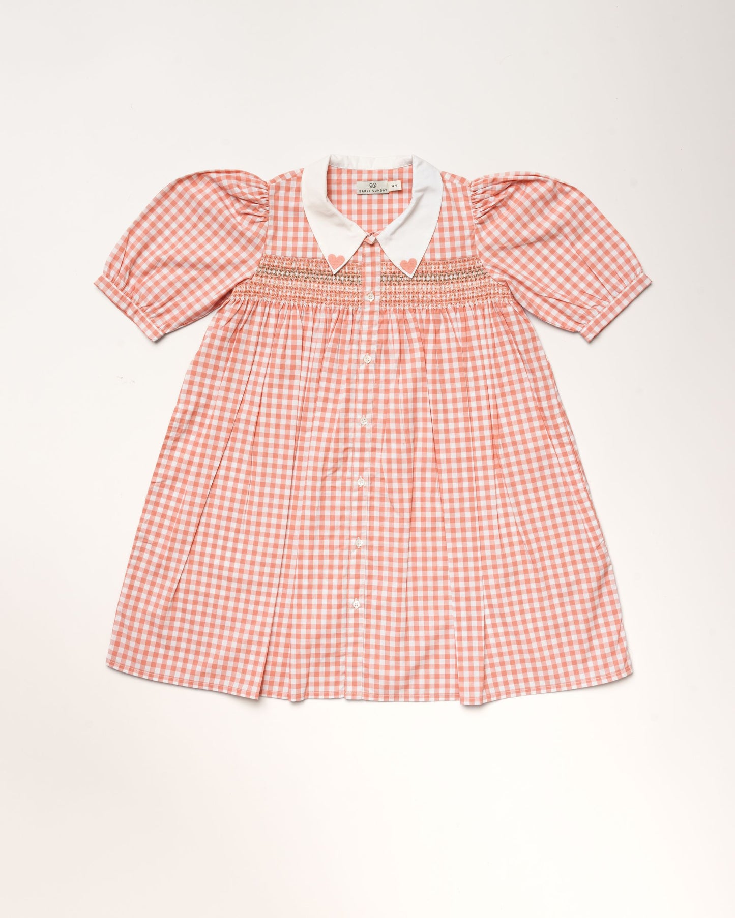 Early Sunday, Jane Dress in Peony color, made from 100% cotton gingham checks fabric, featuring Smocked / embroidered yoke, peter pan collar and puffed short sleeves, shown in a front flat-lay view