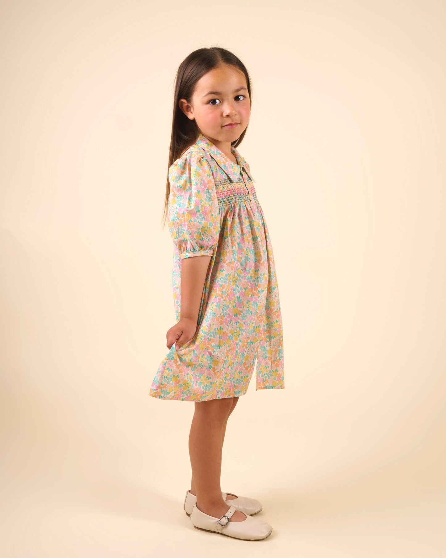 Early Sunday, Jane Dress in Emma Floral Mint color, made from printed 60s cotton cambric fabric, featuring Smocked / embroidered yoke, peter pan collar and puffed short sleeves, shown in a side view of model wearing