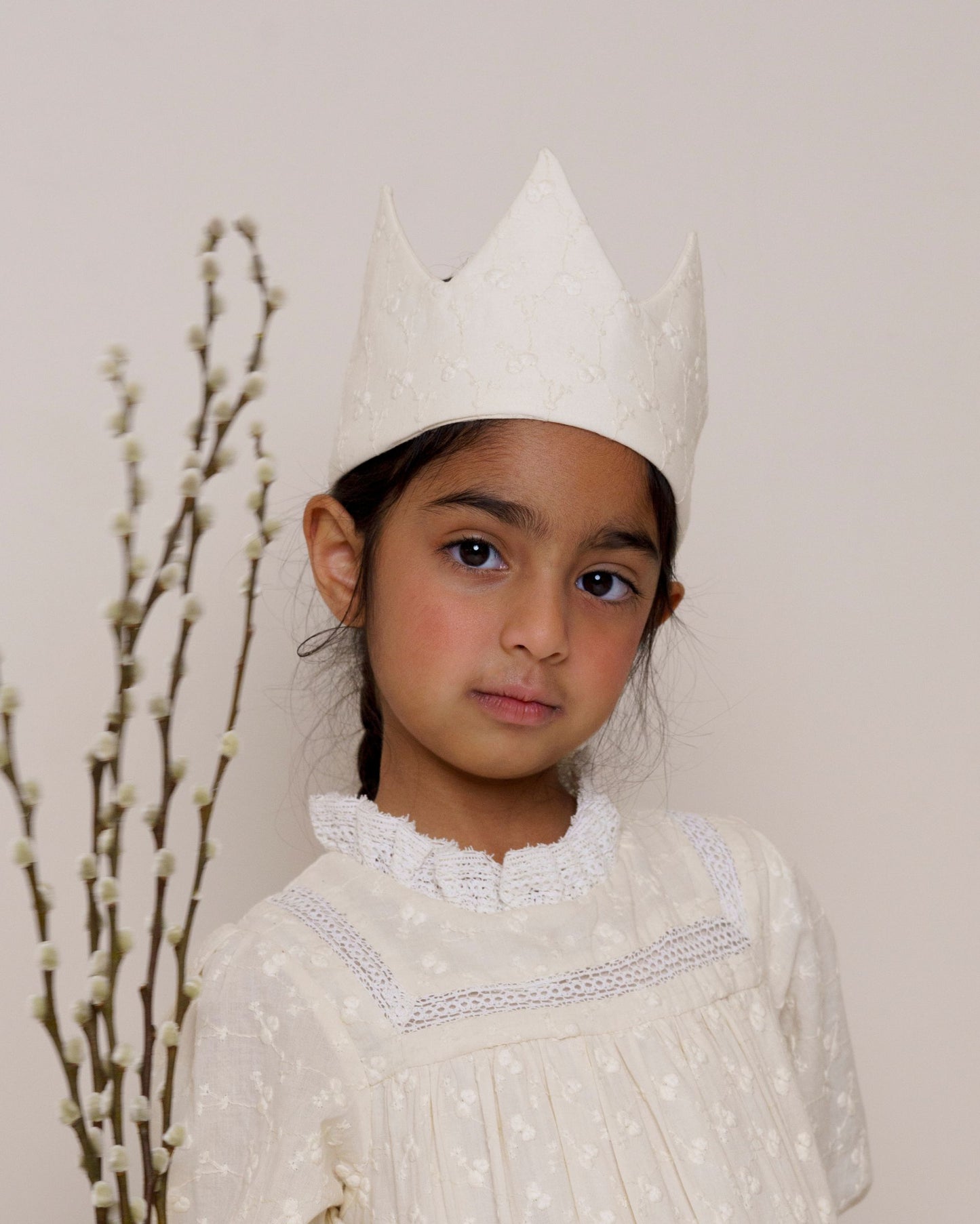 Early Sunday, Kids Crown in Hazelnut cream color, made from All over Embroidered Cotton Cambric fabric, featuring a design with pointed tips & an elasticated band, shown in an angled view of model wearing it