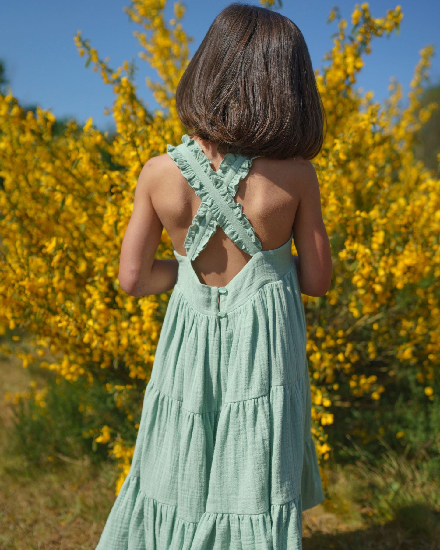 Early Sunday, Laura Dress in Mint color, made from 100% cotton double gauze solid fabric, featuring Crisscross ruffle straps with delicate trim accents and a softly gathered tiered skirt, shown in a back view of model wearing