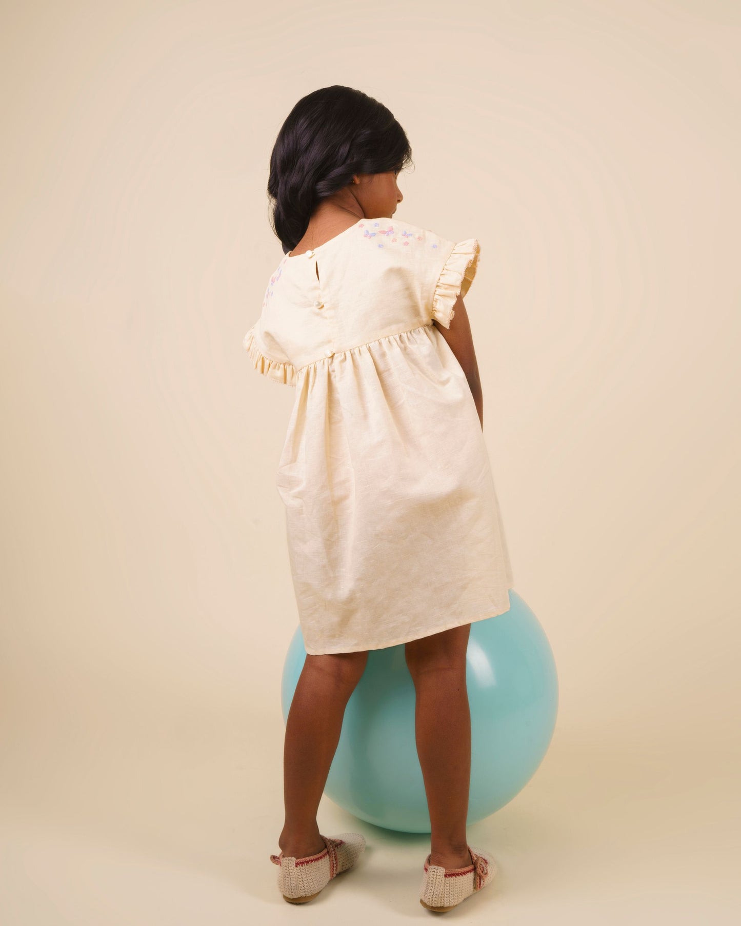 Early Sunday, Lila Dress in Sand color, made from 70% cotton 30% linen solid fabric, featuring Scalloped neckline with contrast stitching, flutter sleeves, gentle gathers, and embroidered hem detail, shown in a back view of model wearing