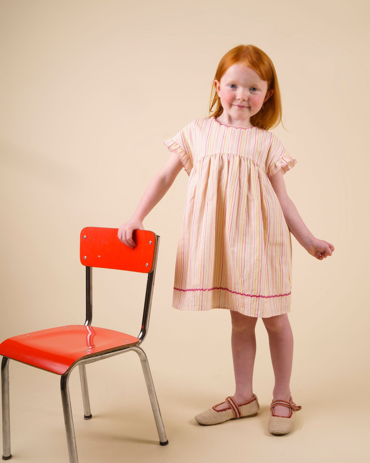 Early Sunday, Lila Dress in Pink Stripes color, made from printed 70% cotton 30% linen fabric, featuring Scalloped neckline with contrast stitching, flutter sleeves, gentle gathers, and embroidered hem detail, shown in a model standing facing front wearing