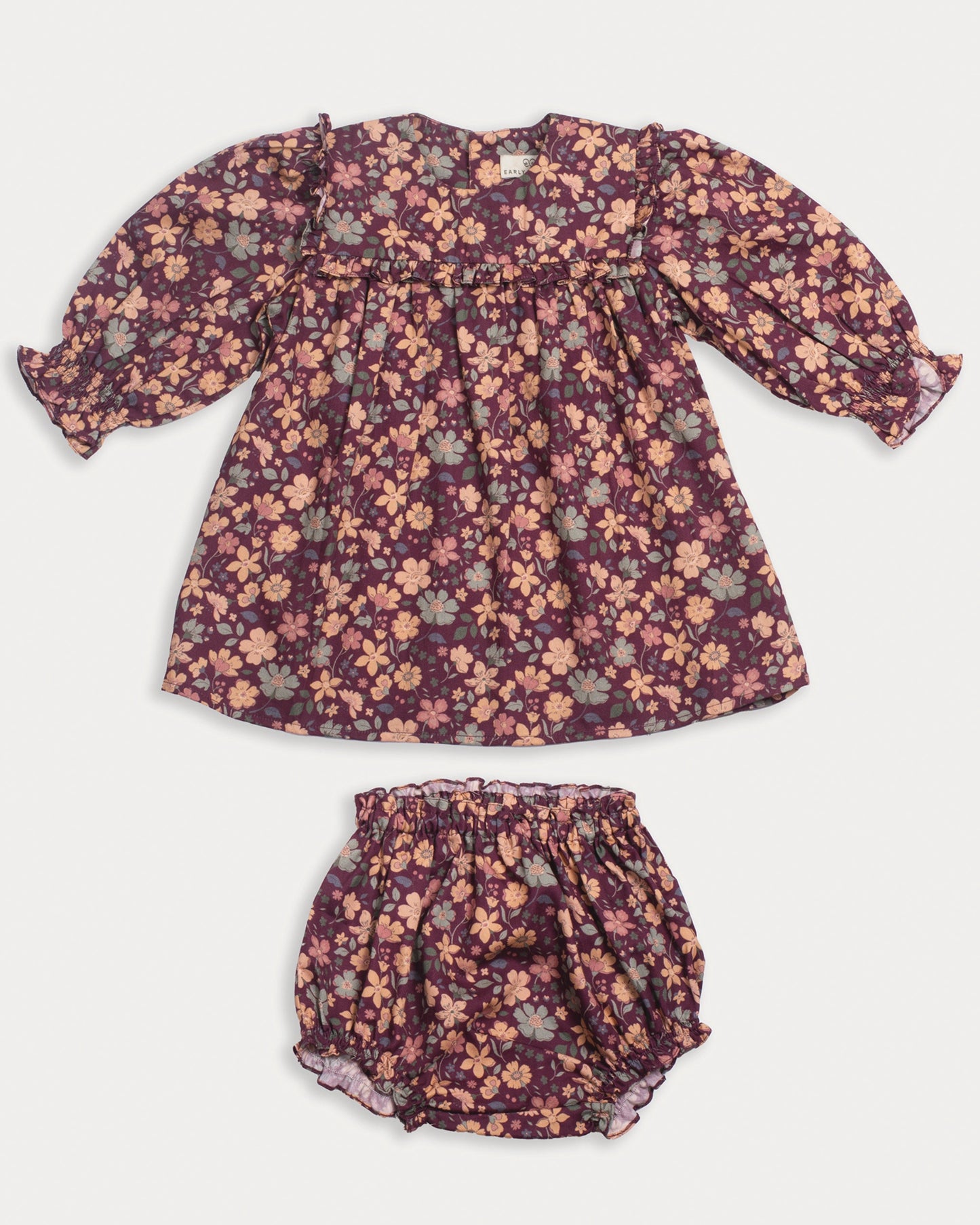Early Sunday, Lottie Baby Dress & Bloomer Set in Garden Flowers Plum, made from Printed Cotton Satin fabric, featuring a frilly neckline & squared frills around the chest, shown in a front flat-lay view