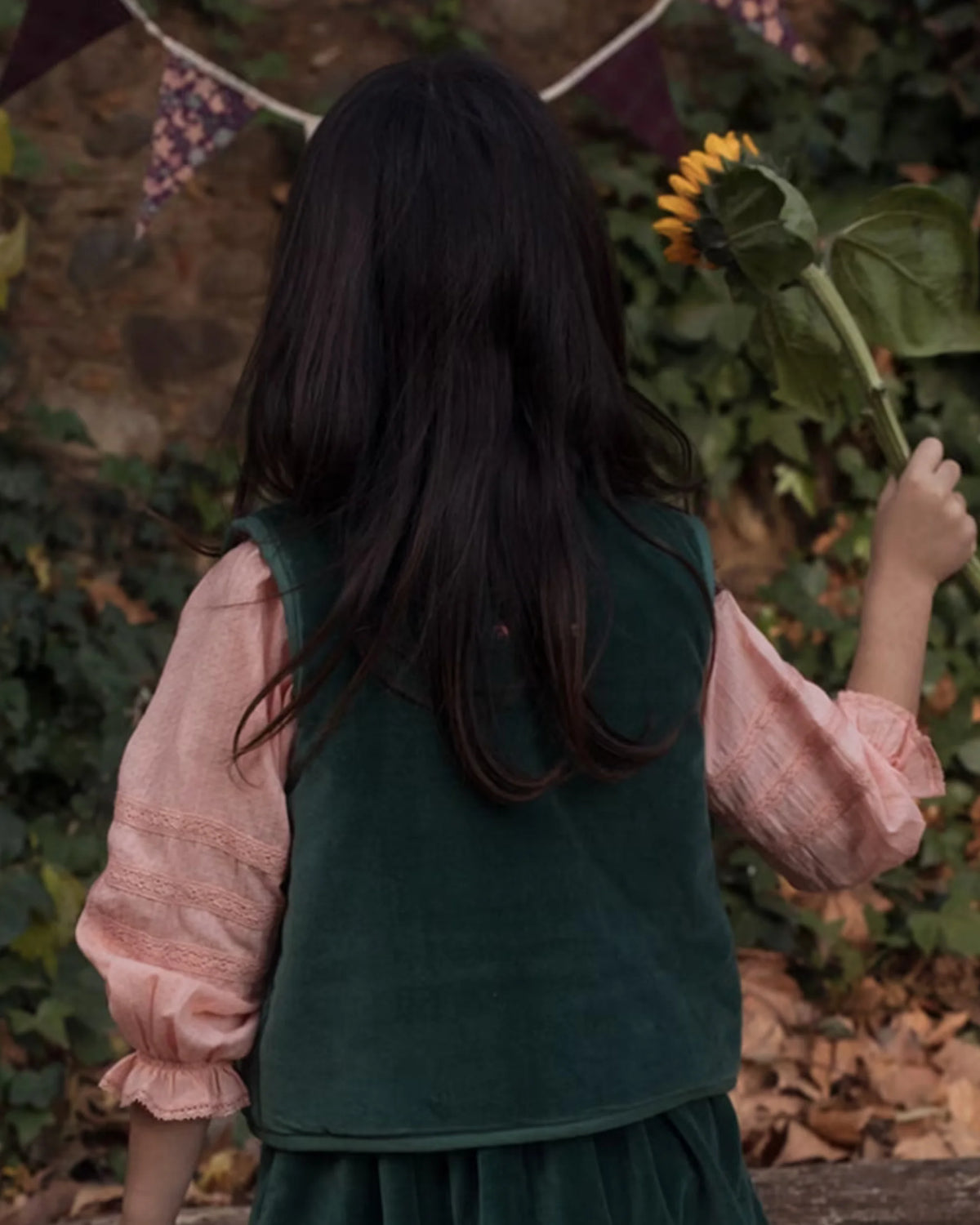 Early Sunday, Lou Vest jacket in Forest Green, made from Cotton Velvet Velour's fabric, featuring sleeveless quilted silhouette with tie-up bow and floral embroidery, shown in a back view of model playing outdoors