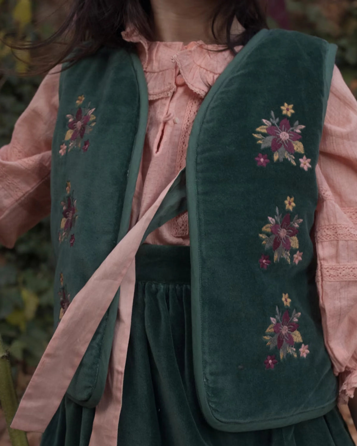 Early Sunday, Lou Vest jacket in Forest Green, made from Cotton Velvet Velour's fabric, featuring sleeveless quilted silhouette with tie-up bow and floral embroidery, shown in a close up shot of model wearing it