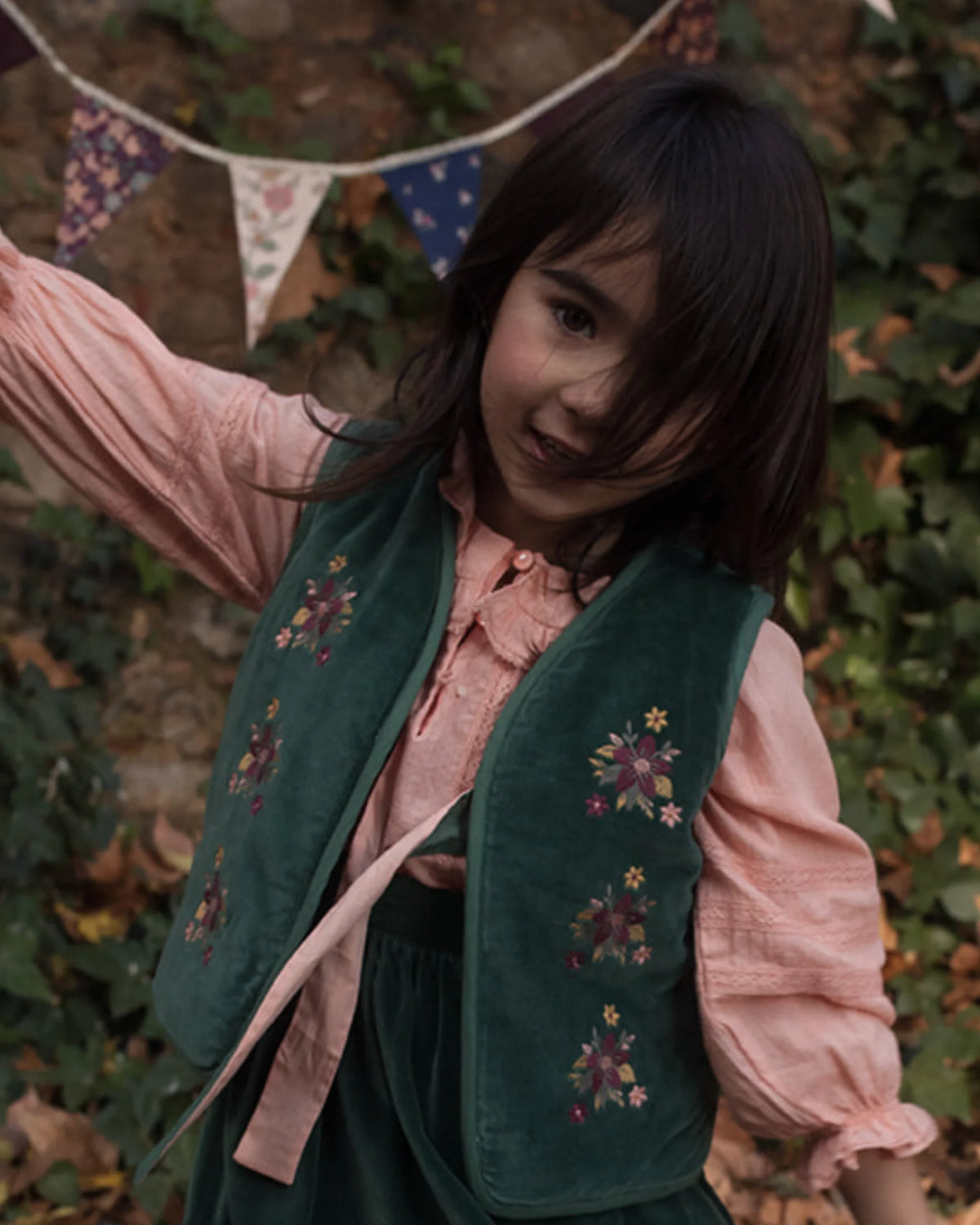 Early Sunday, Lou Vest jacket in Forest Green, made from Cotton Velvet Velour's fabric, featuring sleeveless quilted silhouette with tie-up bow and floral embroidery, shown in a front view of model playing outdoors