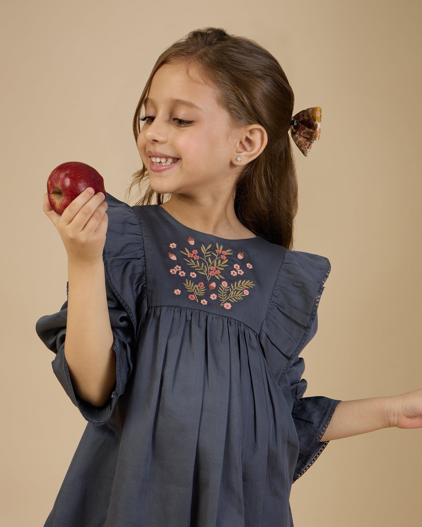 Early Sunday, Margot Dress in Stormy Blue, made from Cotton Satin fabric, featuring classic silhouette with floral embroidery around the yoke and ruffle detailing, shown in a front view of a model wearing it in a studio setting