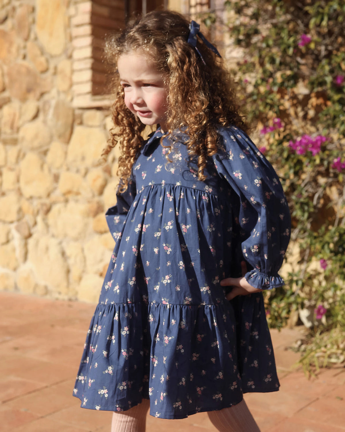 Early Sunday, Marguerite Dress in Ditsy Bouquet Navy, made from Printed Cotton Satin fabric, featuring gathered sleeves with frilled Peter Pan collar, shown in an angled view of a model wearing outdoors