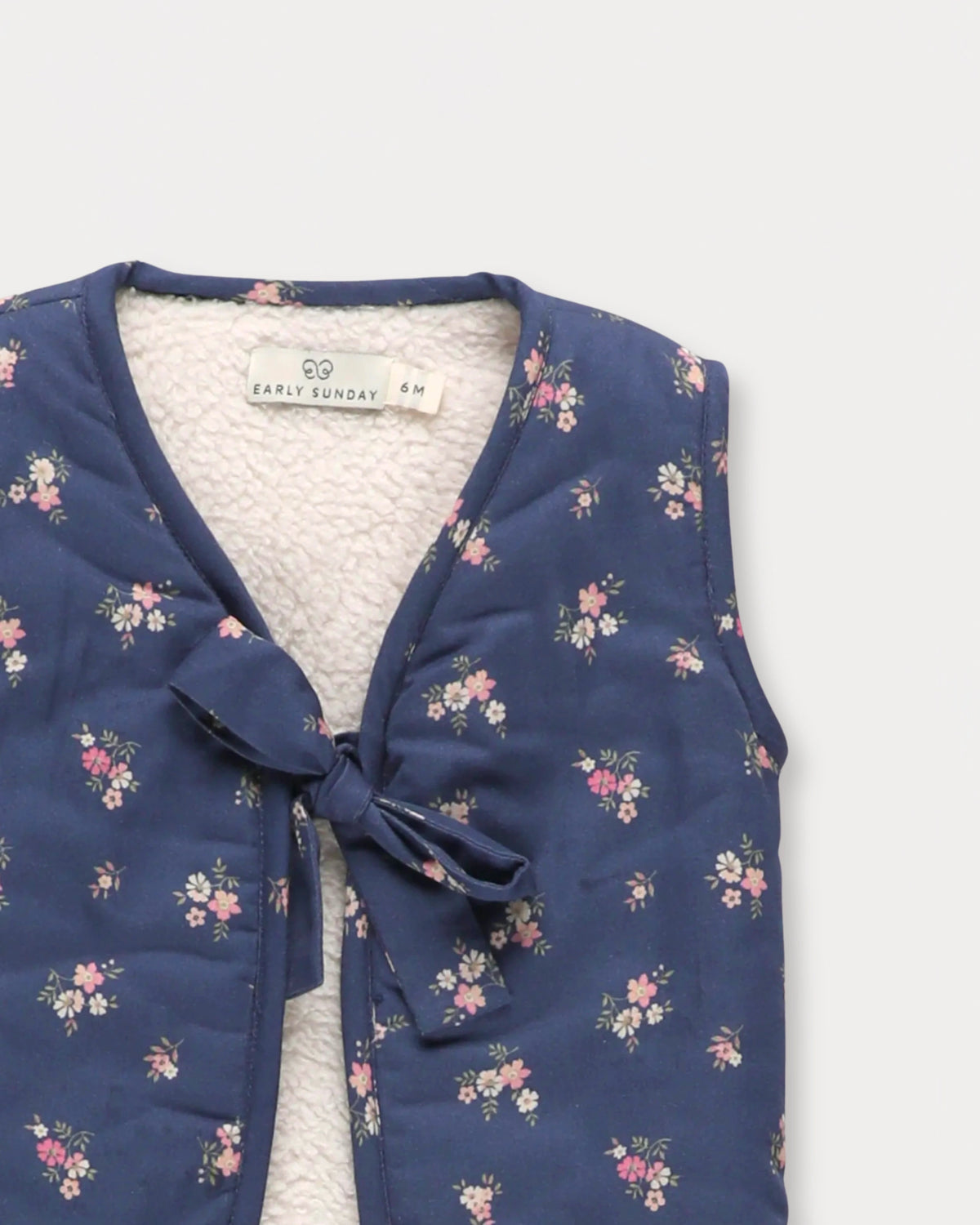 Early Sunday, Milly Baby Quilted Gilet in Ditsy Bouquet Navy, made from Printed Cotton Satin fabric, featuring a sleeveless jacket with a bow tie, shown in a closeup flat-lay view