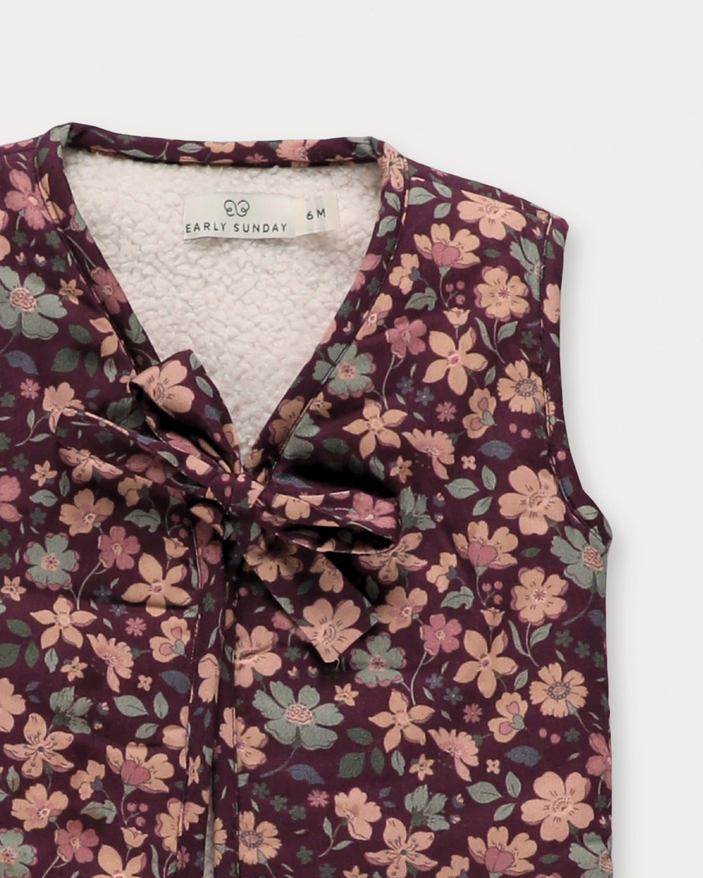 Early Sunday, Milly Baby Quilted Gilet in Garden Flowers Plum, made from Printed Cotton Satin fabric, featuring a sleeveless jacket with a bow tie, shown in a closeup flat-lay view