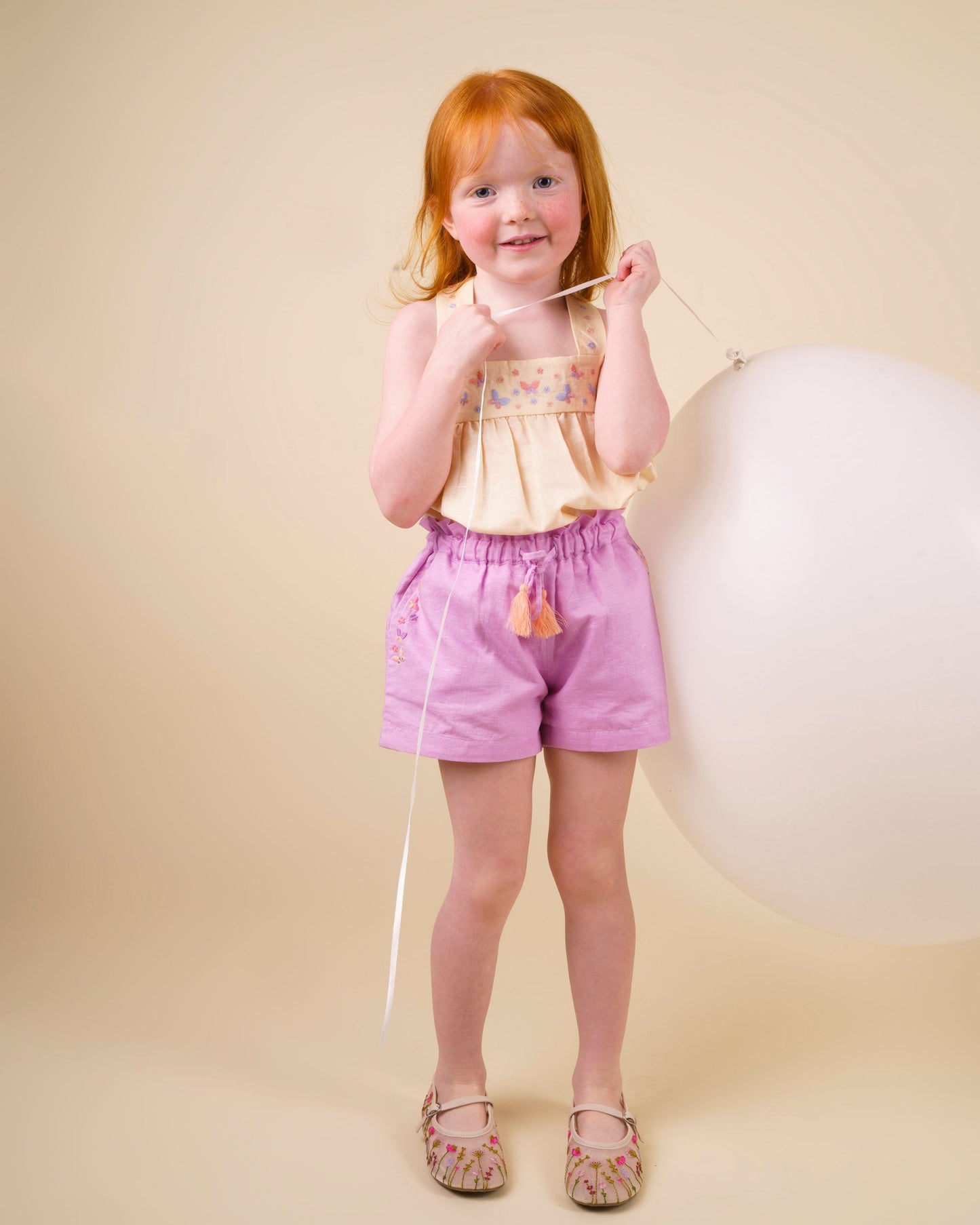Early Sunday, Romy Blouse in Sand color, made from 70% Cotton 30% Linen fabric, featuring a sleeveless design with cross-back straps, a gathered waist & a beautiful embroidery, shown in a front view of a model wearing it in a studio setting