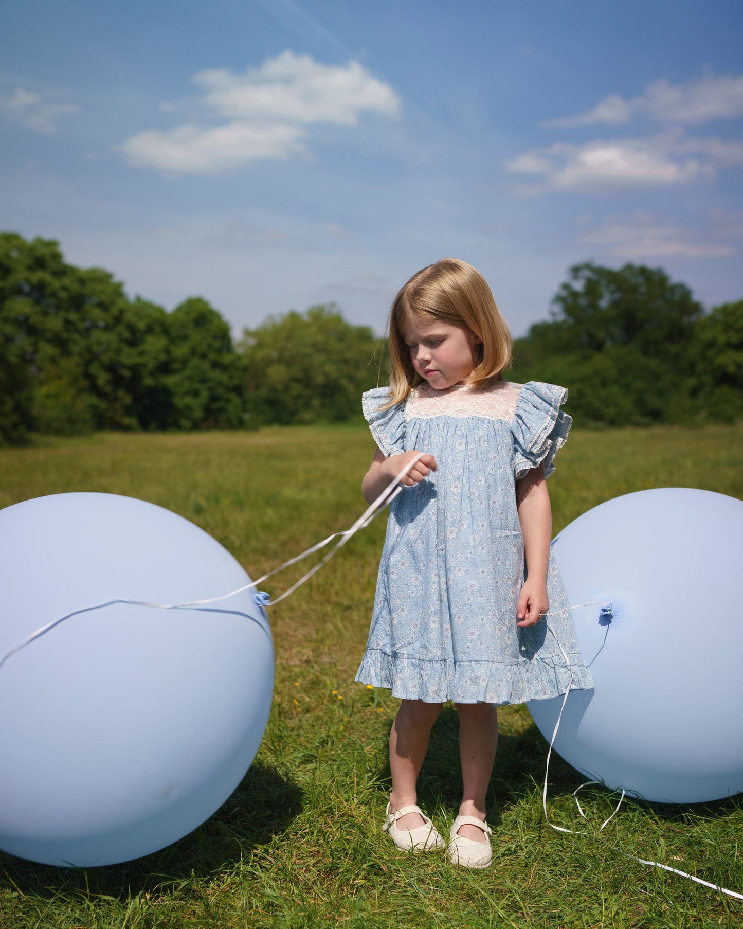 Early Sunday, Sita Dress in Tonal Floral Blue color, made from Printed Cotton Tiny Swiss Dobby fabric, featuring a sleeveless design with wide shoulder straps & a delicate lace yoke, shown in a front view of model wearing outdoors
