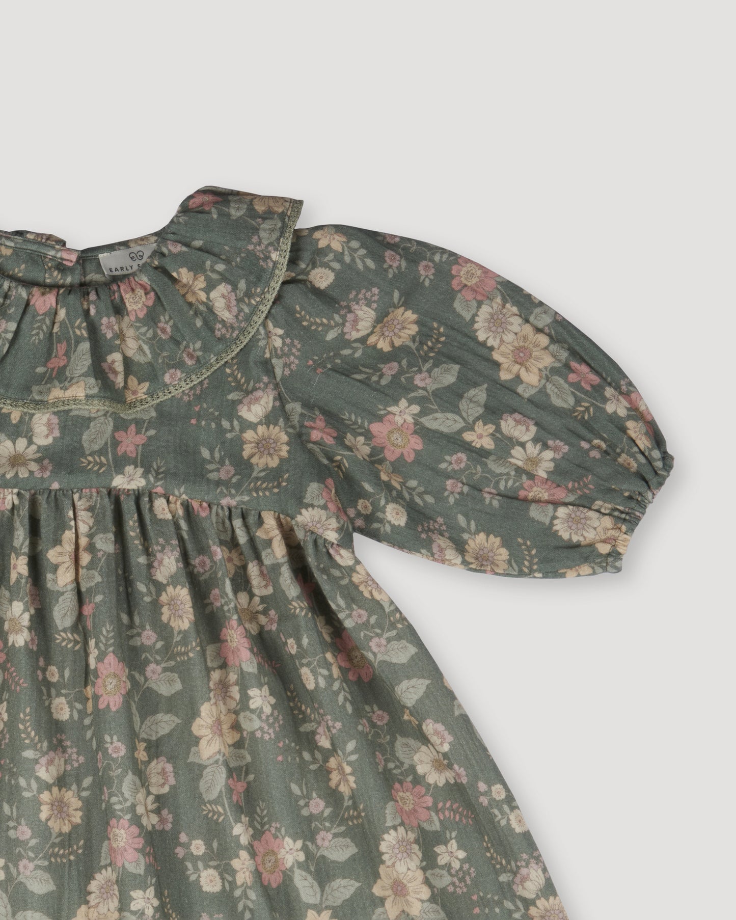 Early Sunday, Suzie Dress in Autumn Bloom Soft Green, made from Printed Cotton Double Gauze fabric, featuring classic silhouette with a ruffle neckline and mother-of-pearl buttons, shown in a closeup flat-lay view