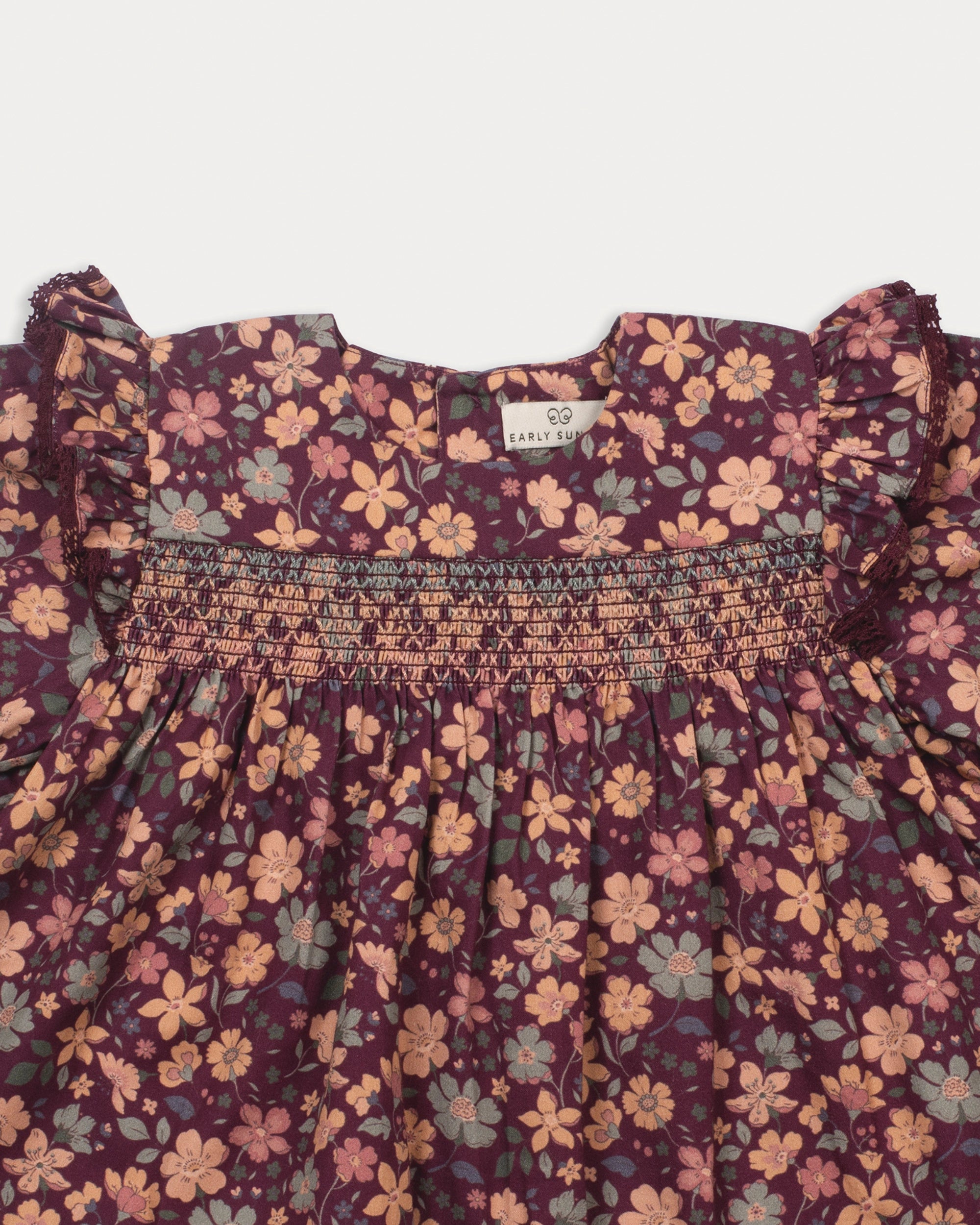 Early Sunday, Ellie Baby Dress & Bloomer Set in Garden Flowers Plum, made from Printed Cotton Satin fabric, featuring frilly sleeves across shoulder & smocked chest, shown in a closeup flat-lay view