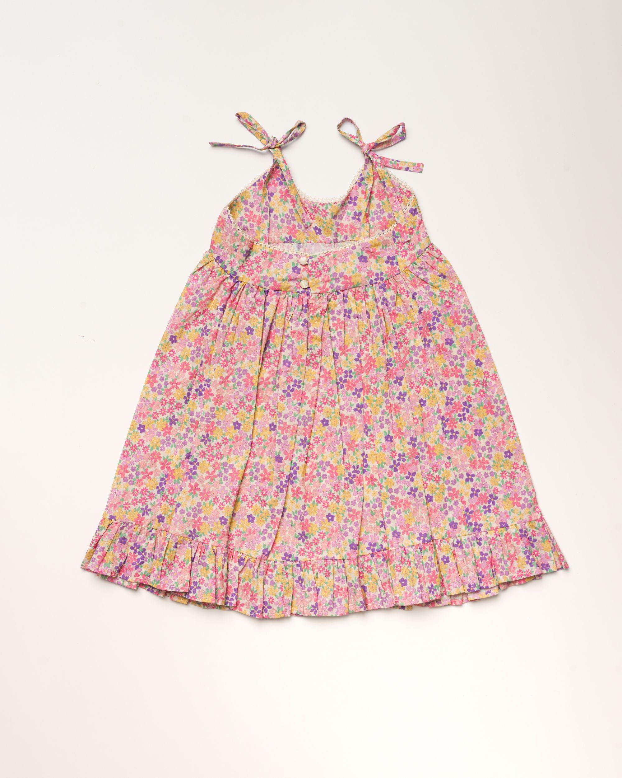 Early Sunday, Alena dress in Emma Floral Purple color, made from organic printed cotton cambric fabric, featuring tie-up shoulder straps, gathered flare, and ruffled hem, shown in a back flat-lay view
