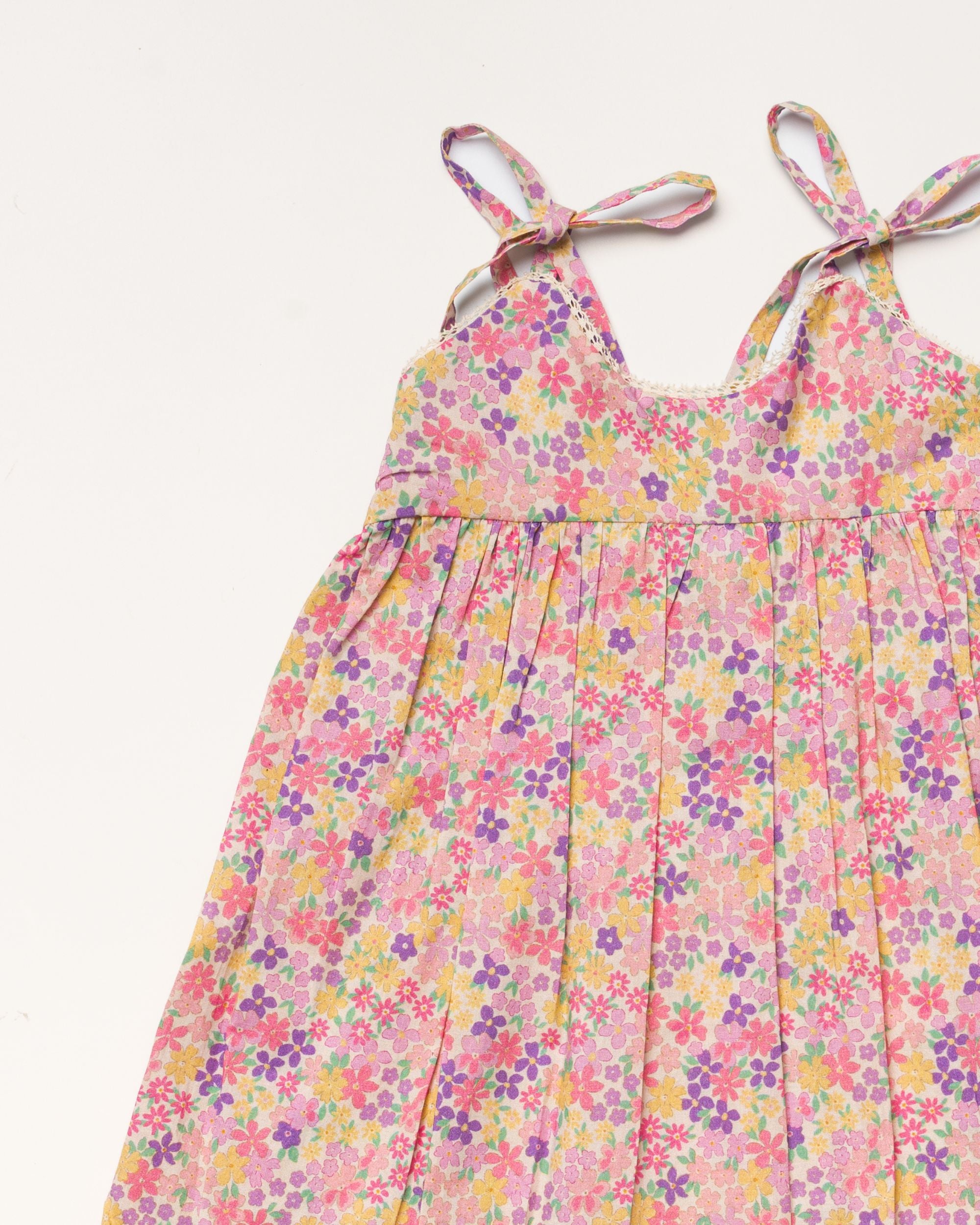 Early Sunday, Alena dress in Emma Floral Purple color, made from organic printed cotton cambric fabric, featuring tie-up shoulder straps, gathered flare, and ruffled hem, shown in a closeup flat-lay view.
