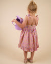 Early Sunday, Alena dress in Emma Floral Purple color, made from organic printed cotton cambric fabric, featuring tie-up shoulder straps, gathered flare, and ruffled hem, shown in a back view of model wearing it.