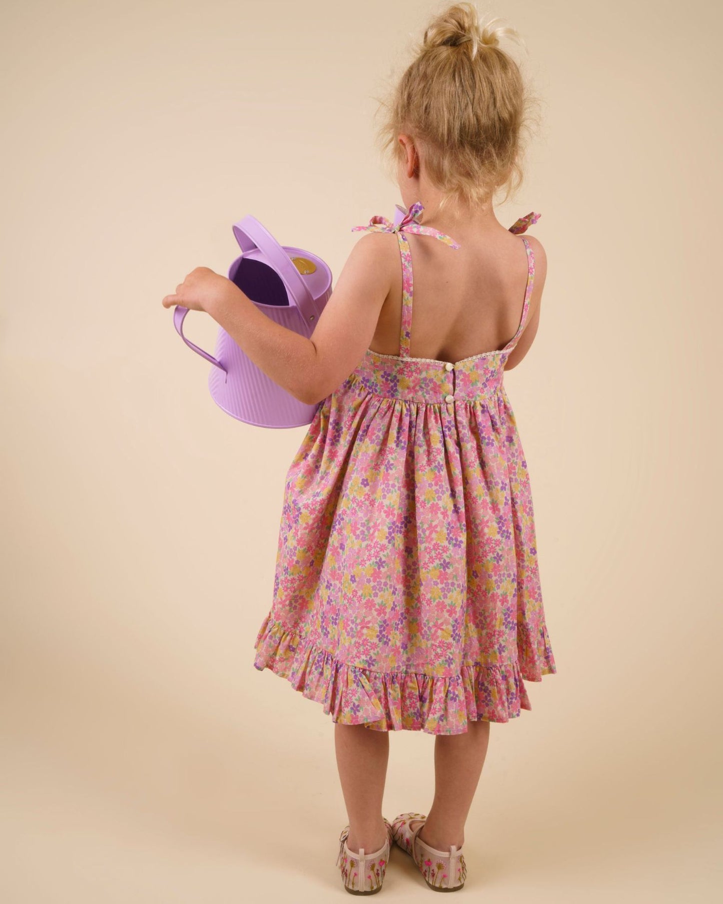 Early Sunday, Alena dress in Emma Floral Purple color, made from organic printed cotton cambric fabric, featuring tie-up shoulder straps, gathered flare, and ruffled hem, shown in a back view of model wearing it.