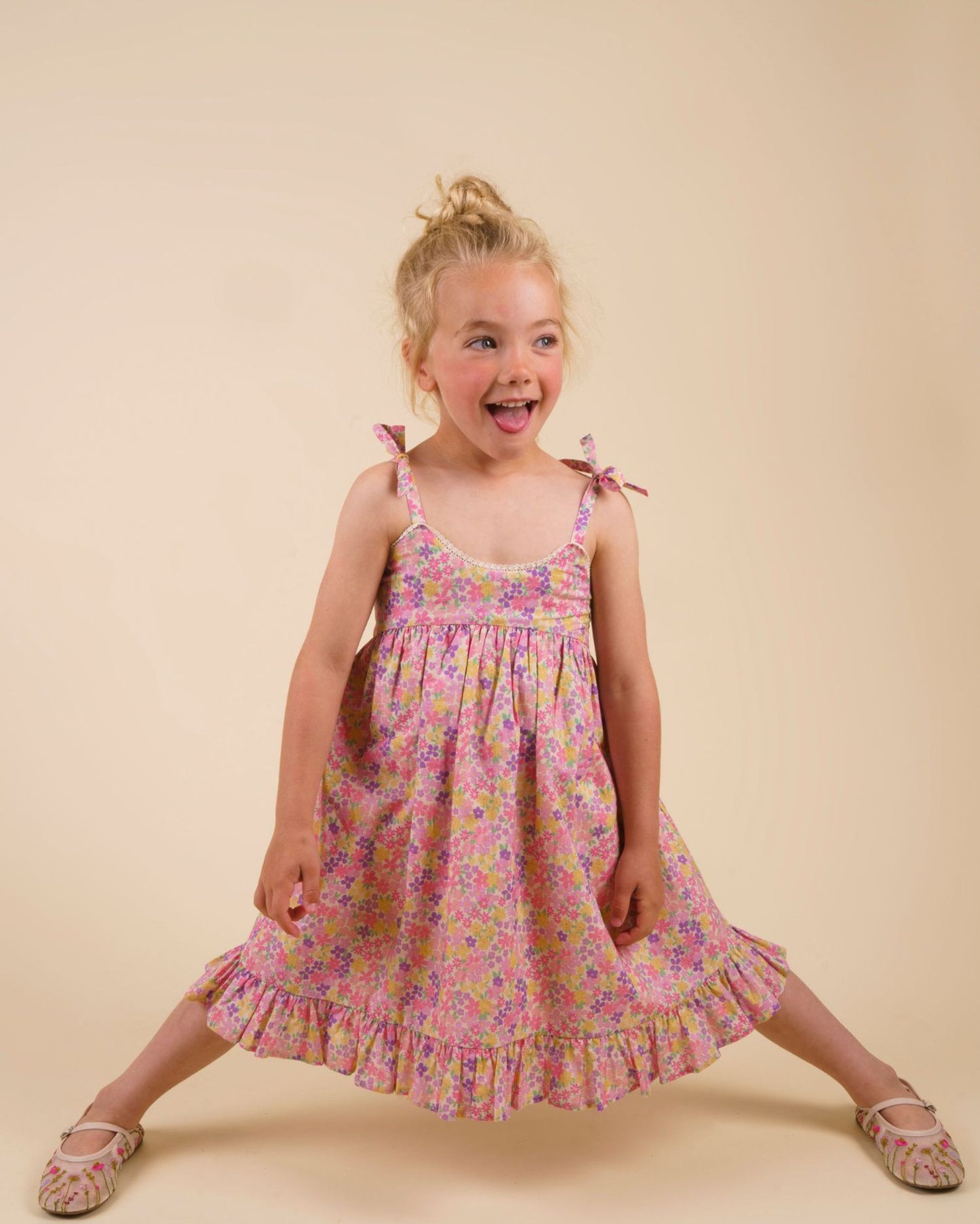 Early Sunday, Alena dress in Emma Floral Purple color, made from organic printed cotton cambric fabric, featuring tie-up shoulder straps, gathered flare, and ruffled hem, shown in a model standing facing front wearing it.
