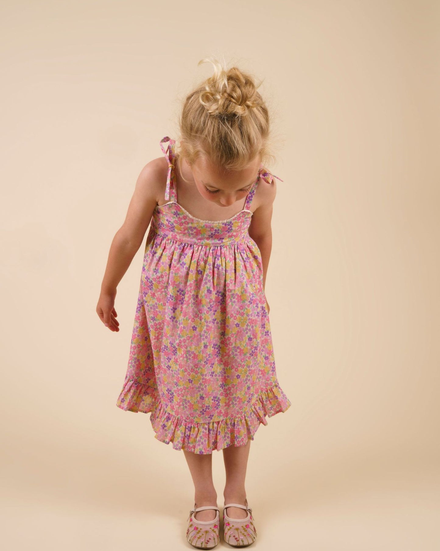 Early Sunday, Alena dress in Emma Floral Purple color, made from organic printed cotton cambric fabric, featuring tie-up shoulder straps, gathered flare, and ruffled hem, shown in a front view of model wearing it.