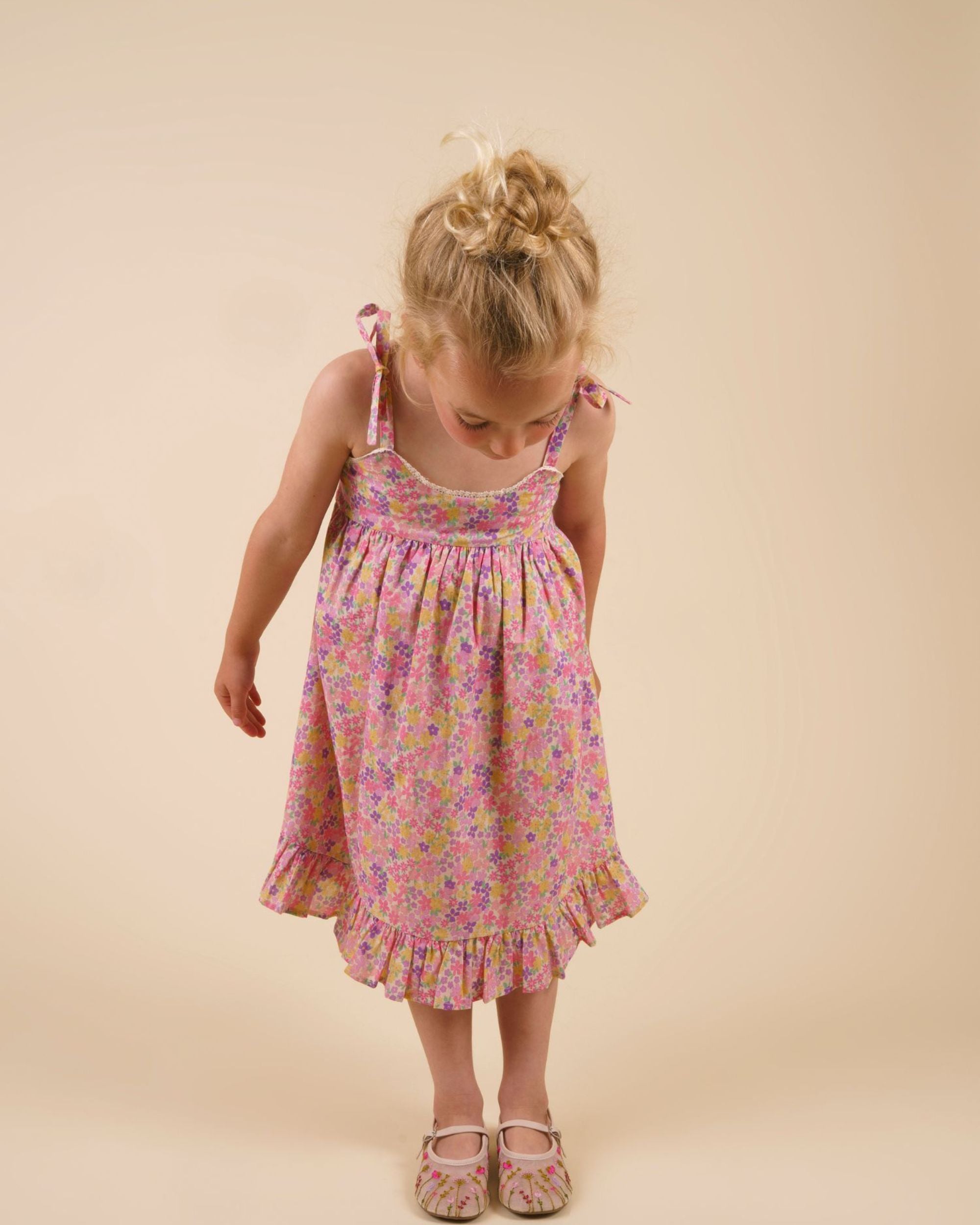 Early Sunday, Alena dress in Emma Floral Purple color, made from organic printed cotton cambric fabric, featuring tie-up shoulder straps, gathered flare, and ruffled hem, shown in a front view of model wearing it.