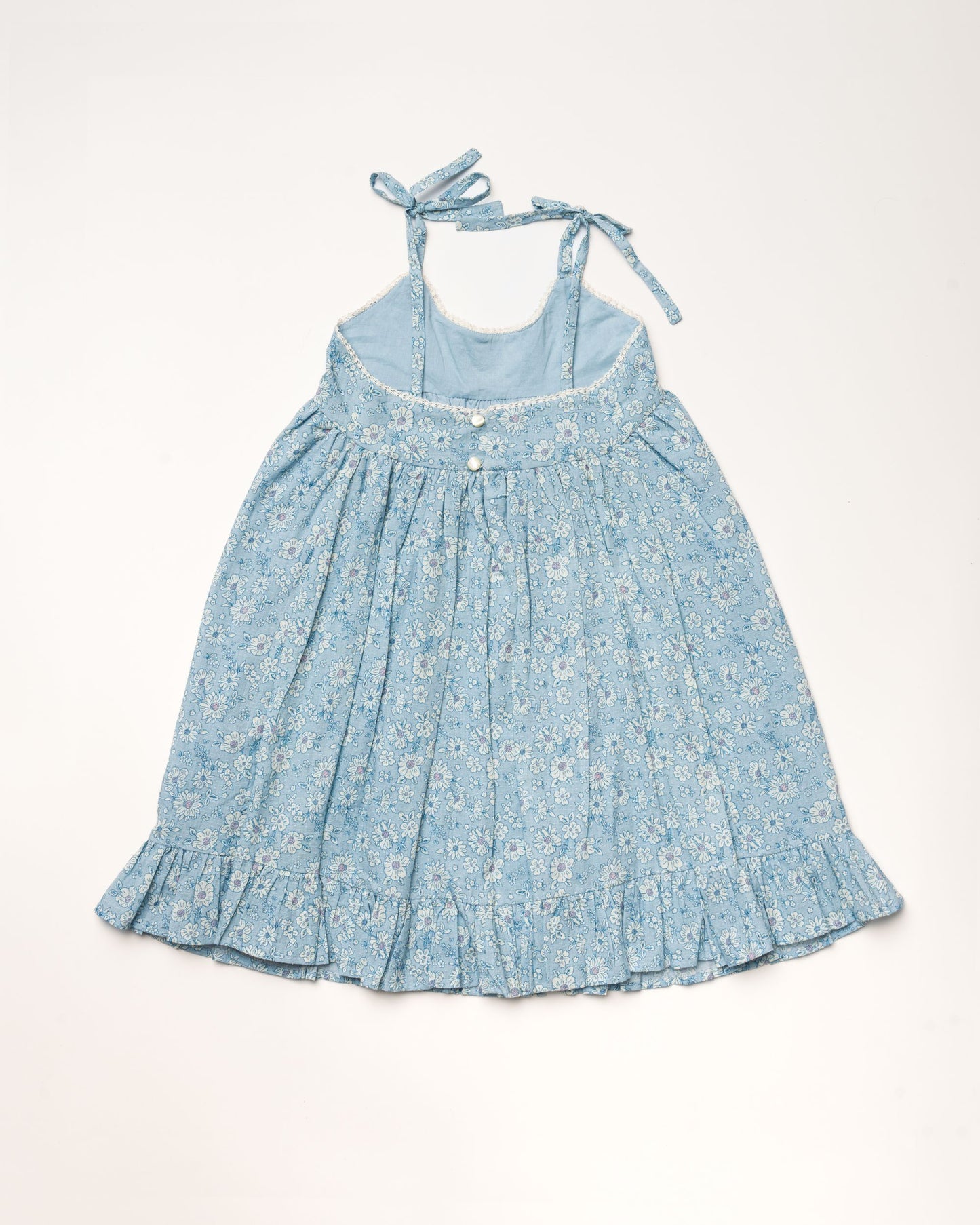 Early Sunday, Alena dress in Tonal Floral Blue color, made from printed cotton tiny swiss dobby fabric, featuring tie-up shoulder straps, gathered flare, and ruffled hem, shown in a back flat-lay view.
