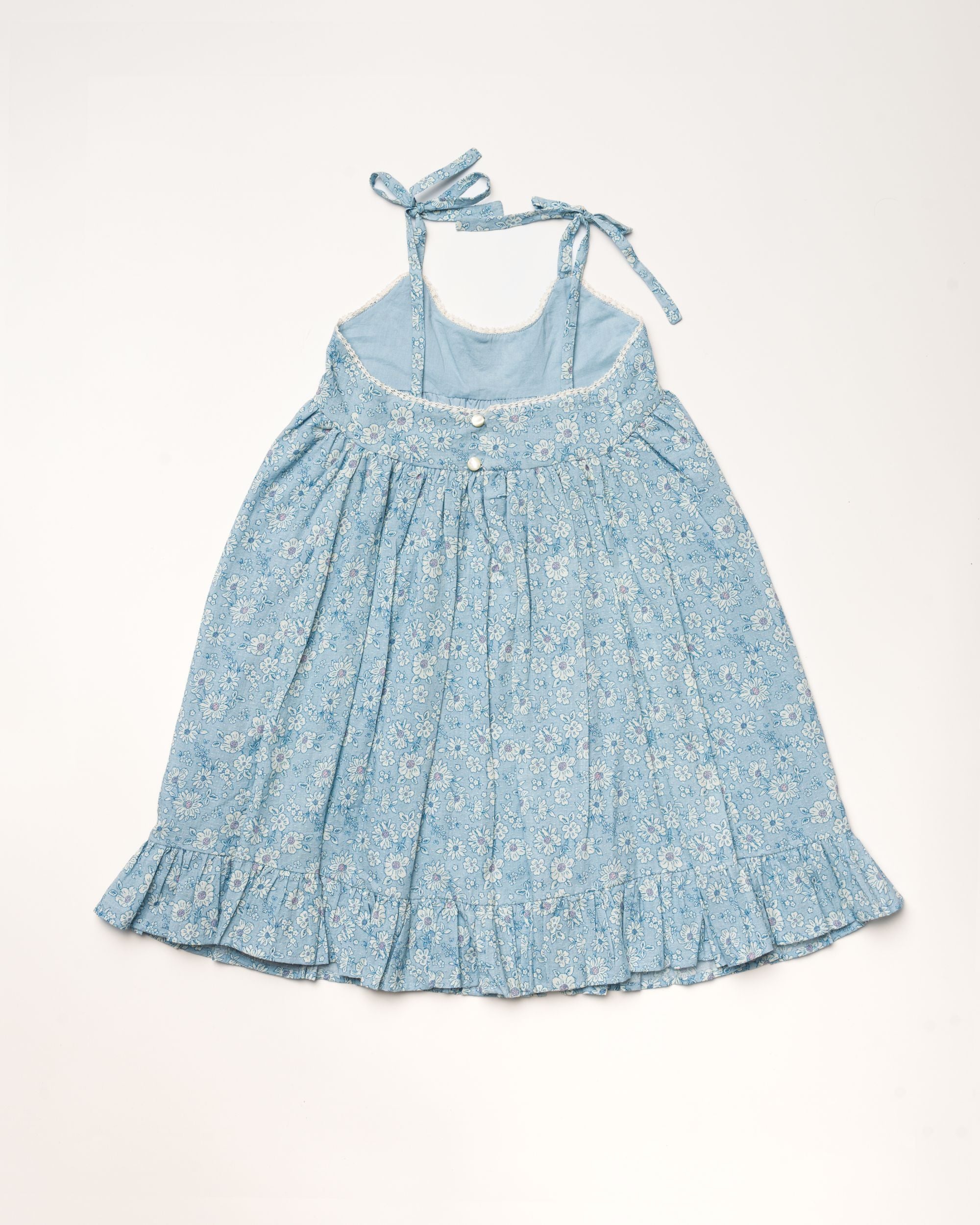 Early Sunday, Alena dress in Tonal Floral Blue color, made from printed cotton tiny swiss dobby fabric, featuring tie-up shoulder straps, gathered flare, and ruffled hem, shown in a back flat-lay view.