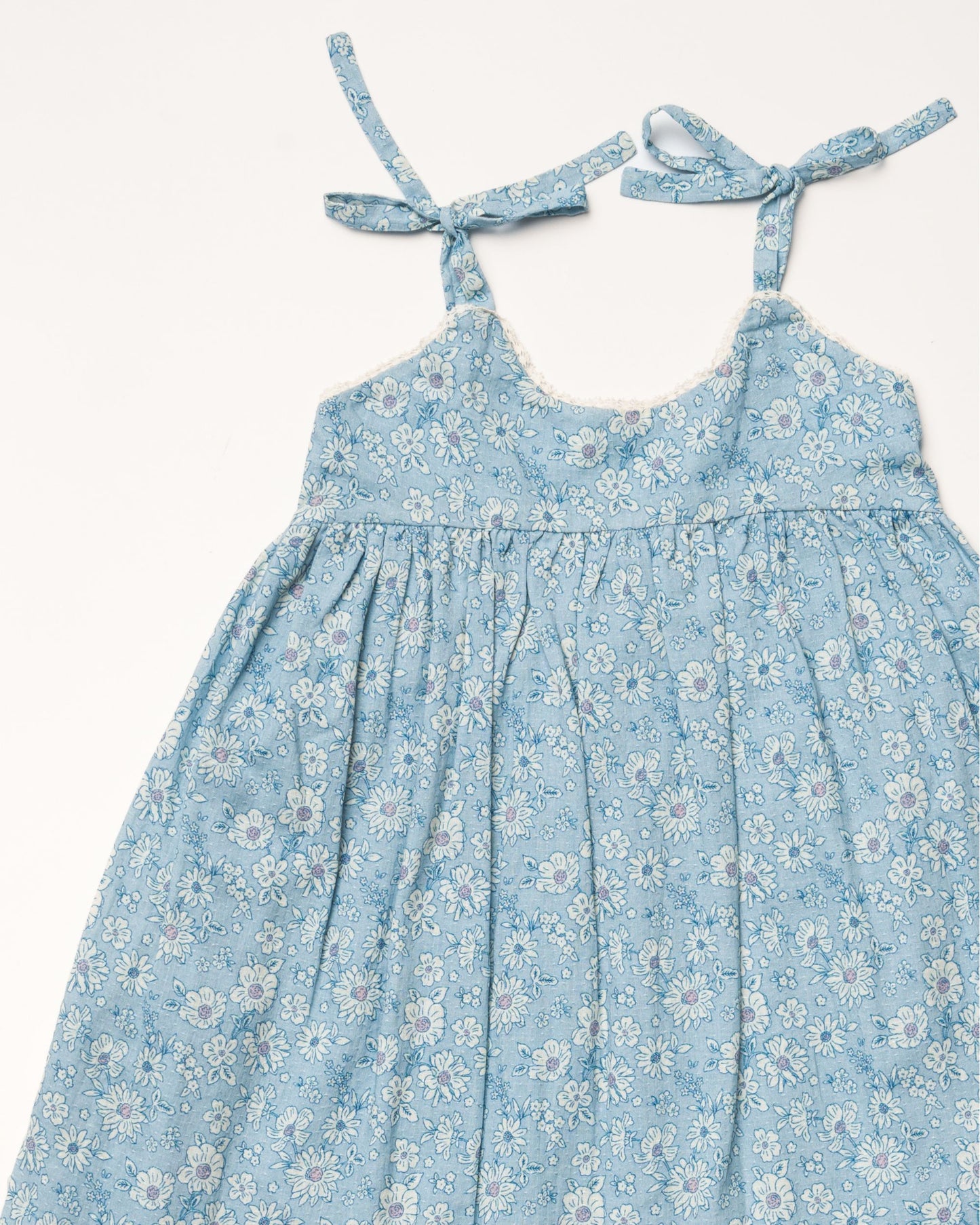 Early Sunday, Alena dress in Tonal Floral Blue color, made from printed cotton tiny swiss dobby fabric, featuring tie-up shoulder straps, gathered flare, and ruffled hem, shown in a closeup flat-lay view.