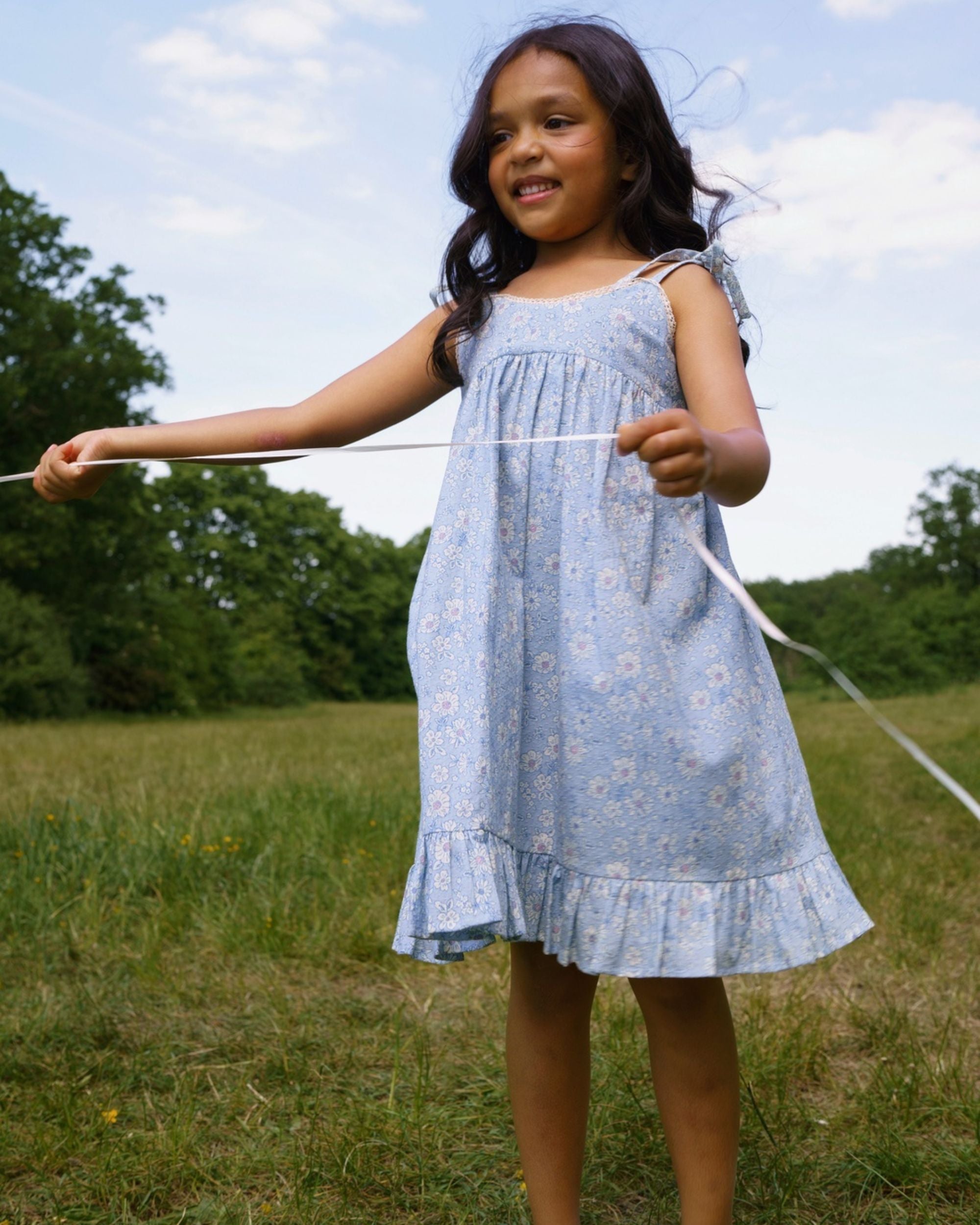 Early Sunday, Alena dress in Tonal Floral Blue color, made from printed cotton tiny swiss dobby fabric, featuring tie-up shoulder straps, gathered flare, and ruffled hem, shown in a model in motion.