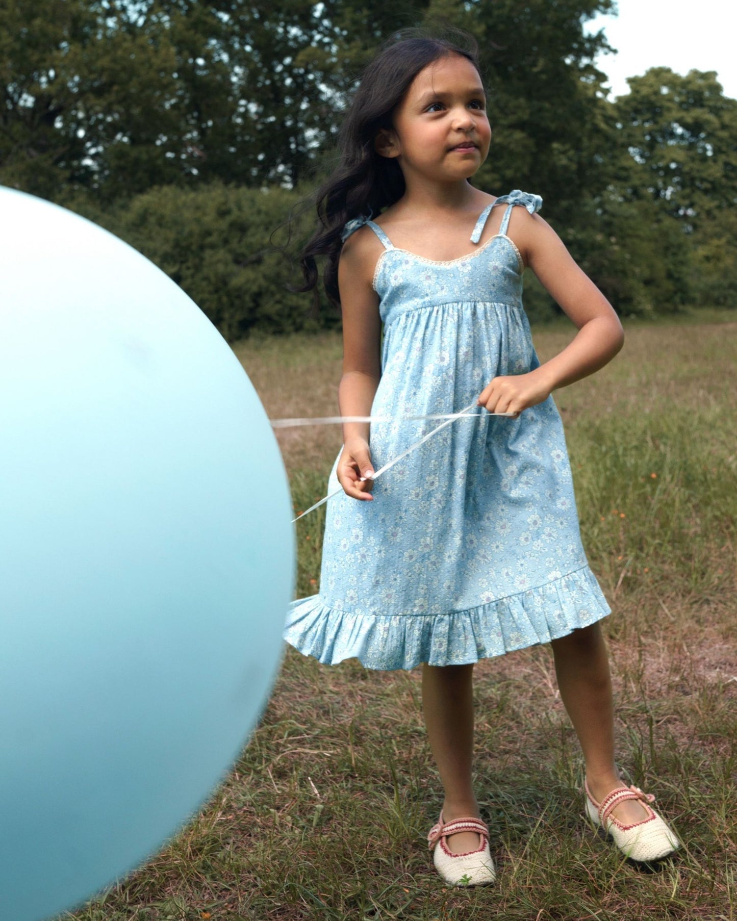 Early Sunday, Alena dress in Tonal Floral Blue color, made from printed cotton tiny swiss dobby fabric, featuring tie-up shoulder straps, gathered flare, and ruffled hem, shown in a front view of model wearing.