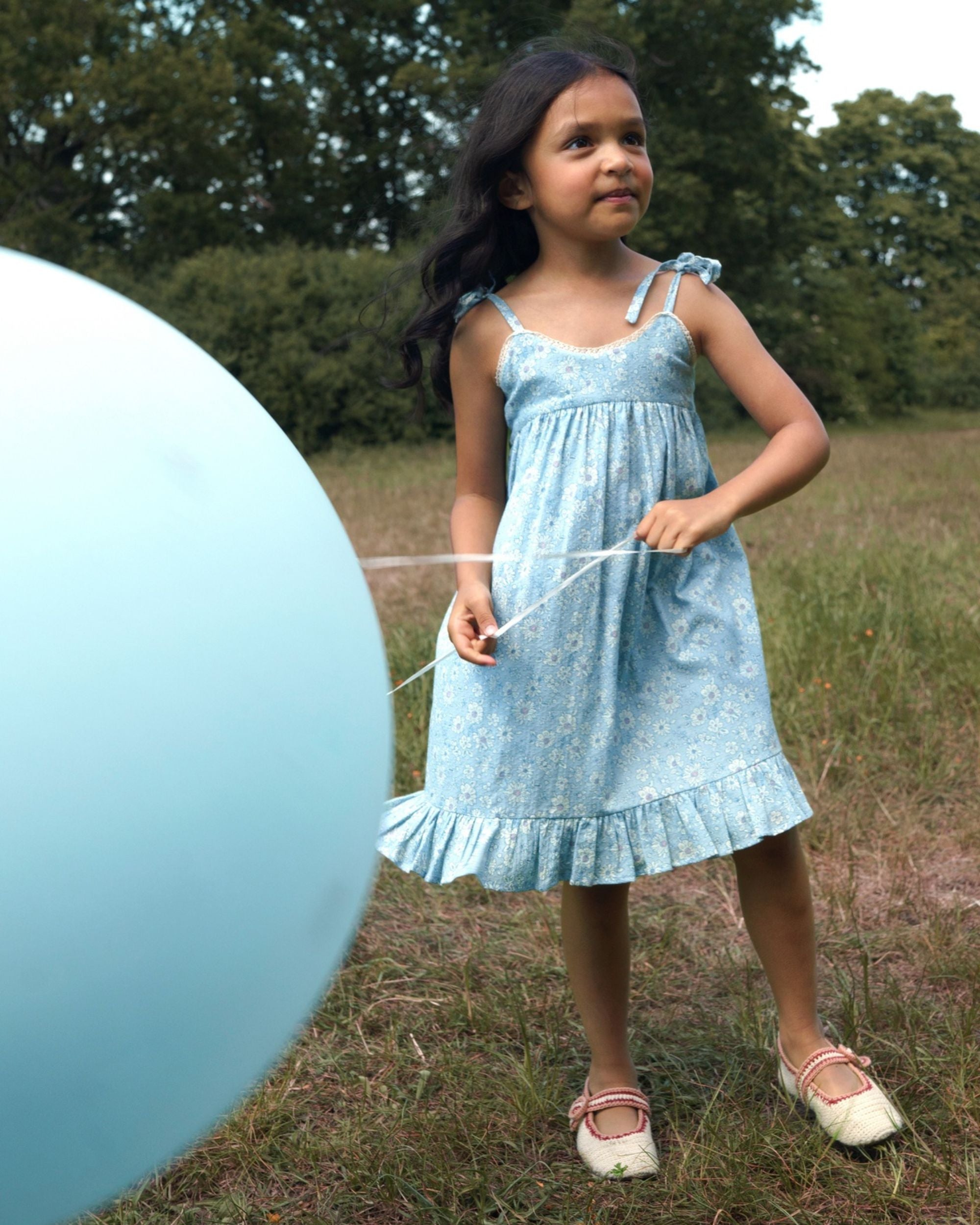 Early Sunday, Alena dress in Tonal Floral Blue color, made from printed cotton tiny swiss dobby fabric, featuring tie-up shoulder straps, gathered flare, and ruffled hem, shown in a front view of model wearing.