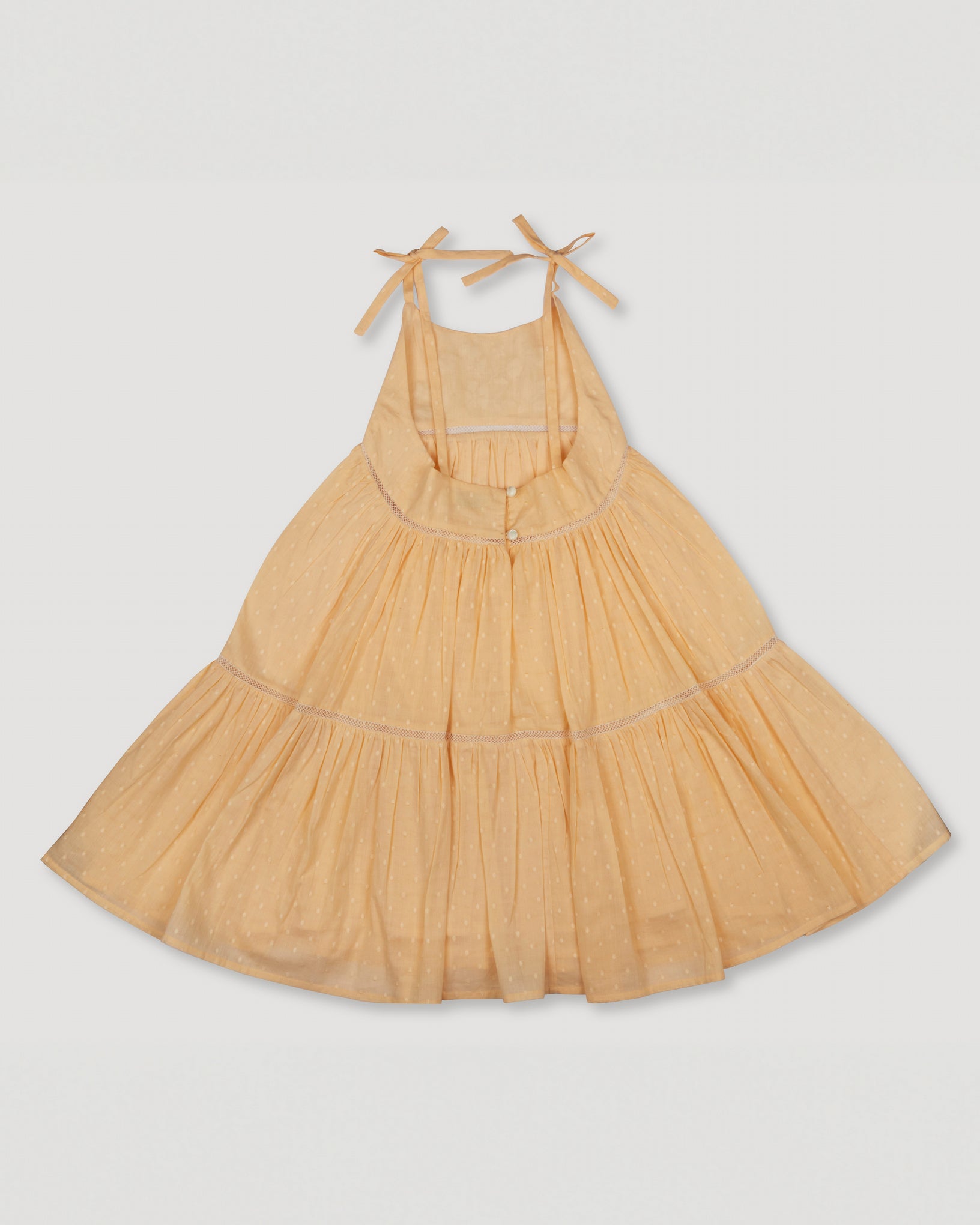 Early Sunday, Alice Dress in Vanilla, made from Cotton Dobby Swiss Dots fabric, featuring sleeveless with adjustable tie-up straps and a tiered silhouette, shown in a back flat-lay view