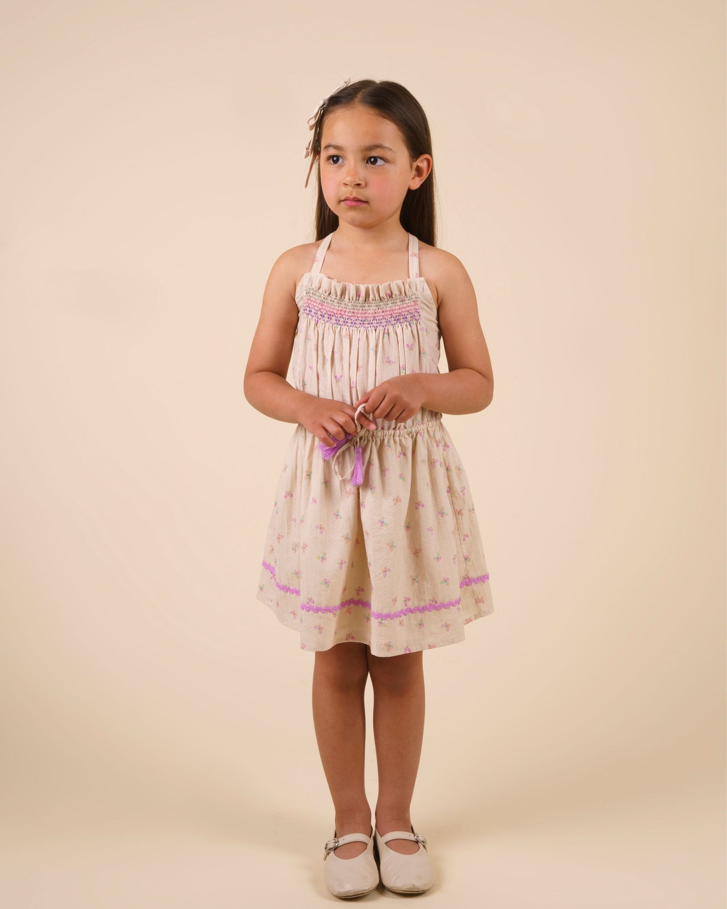 Early Sunday, Alisson Dress in butterfly print color, made from printed cotton tiny swiss dobby fabric, featuring smocked bodice, adjustable shoulder straps, sweet tassel accents, and a flowy gathered skirt, shown in a model standing facing front wearing.