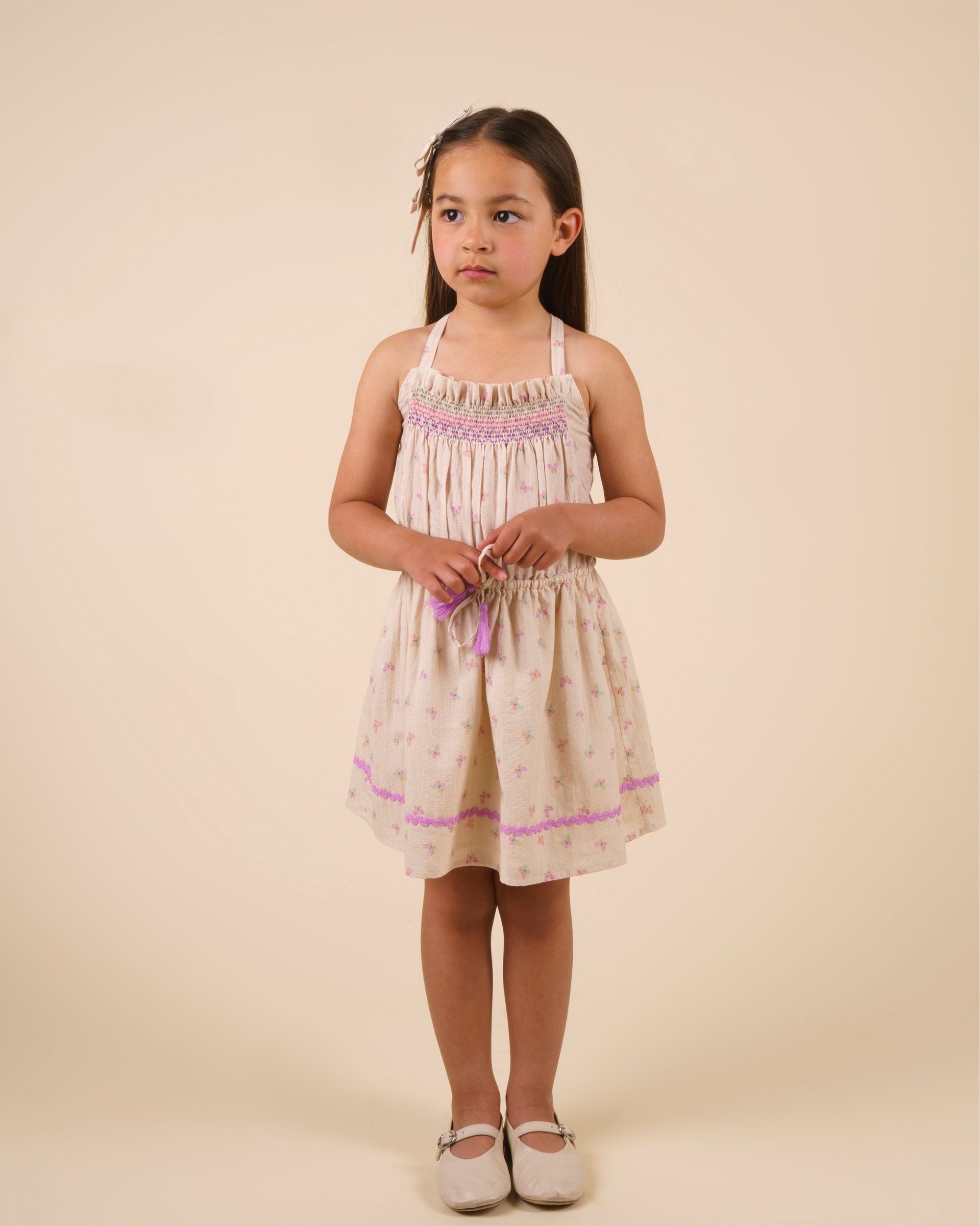 Early Sunday, Alisson Dress in butterfly print color, made from printed cotton tiny swiss dobby fabric, featuring smocked bodice, adjustable shoulder straps, sweet tassel accents, and a flowy gathered skirt, shown in a model standing facing front wearing.