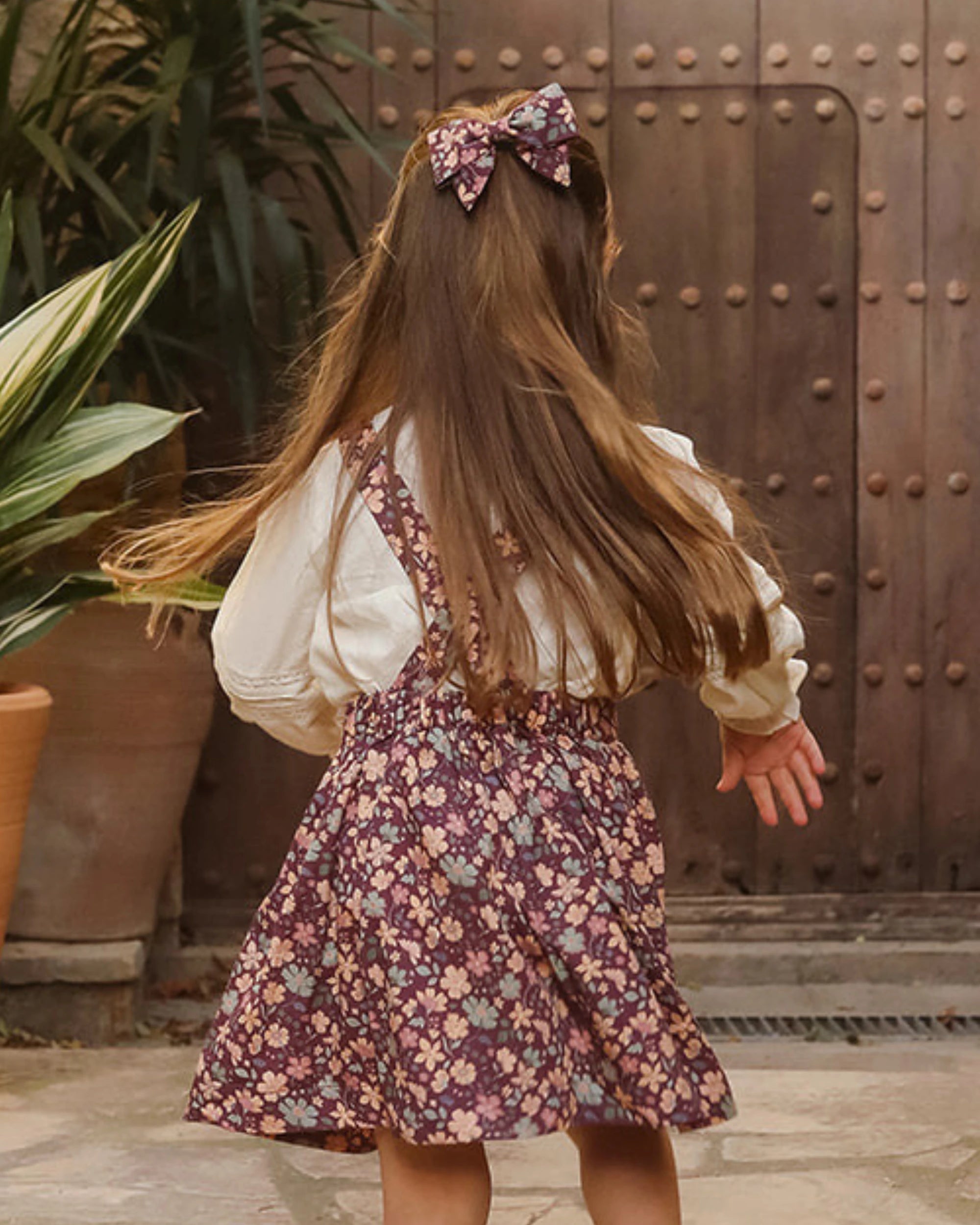 Early Sunday, Annie Skirt in Garden Flowers Plum, made from Printed Cotton Satin fabric, featuring an A-line silhouette with adjustable cross-back straps and soft gathers, shown in a back view of model wearing outdoors