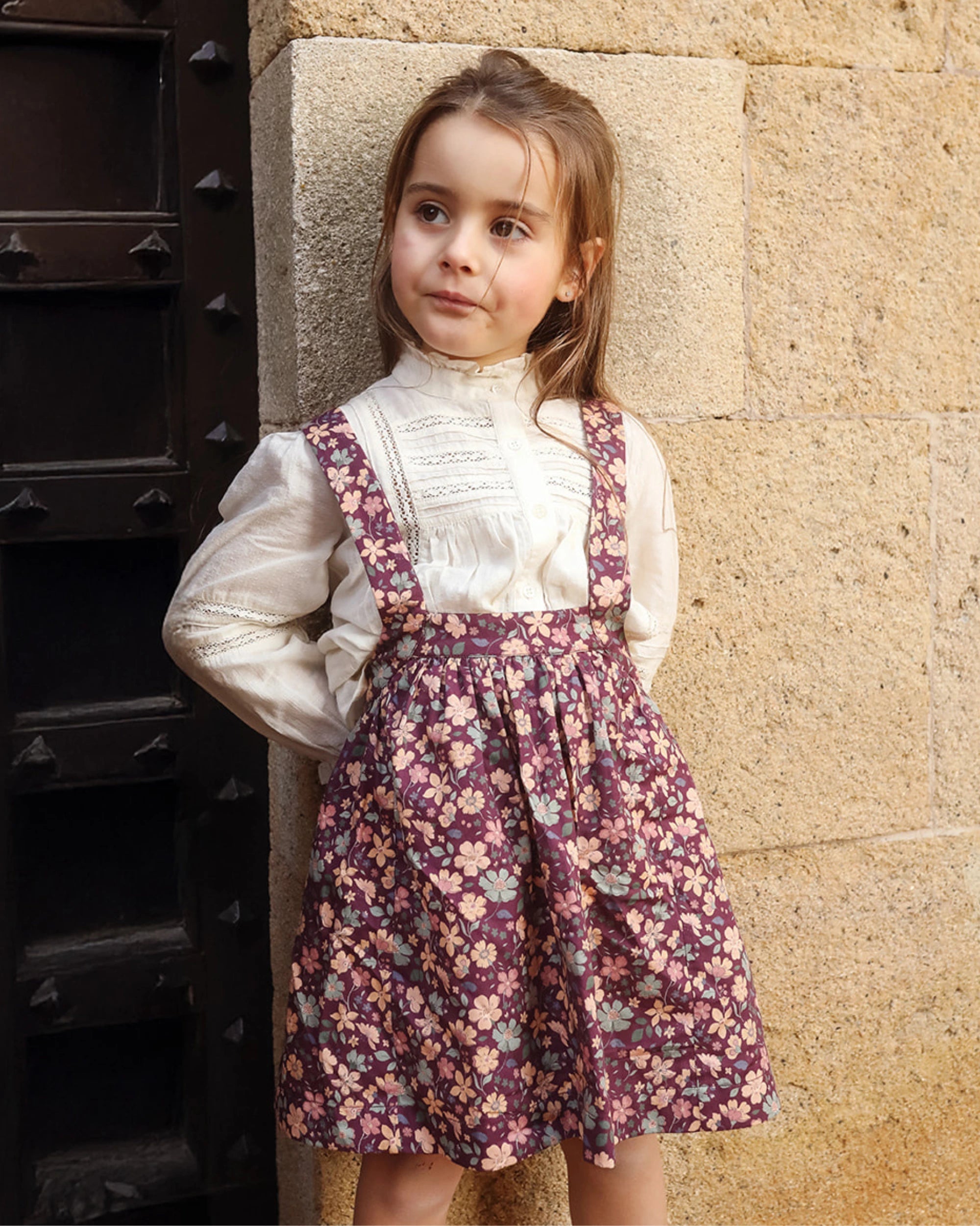 Early Sunday, Annie Skirt in Garden Flowers Plum, made from Printed Cotton Satin fabric, featuring an A-line silhouette with adjustable cross-back straps and soft gathers, shown in a front view of model wearing outdoors