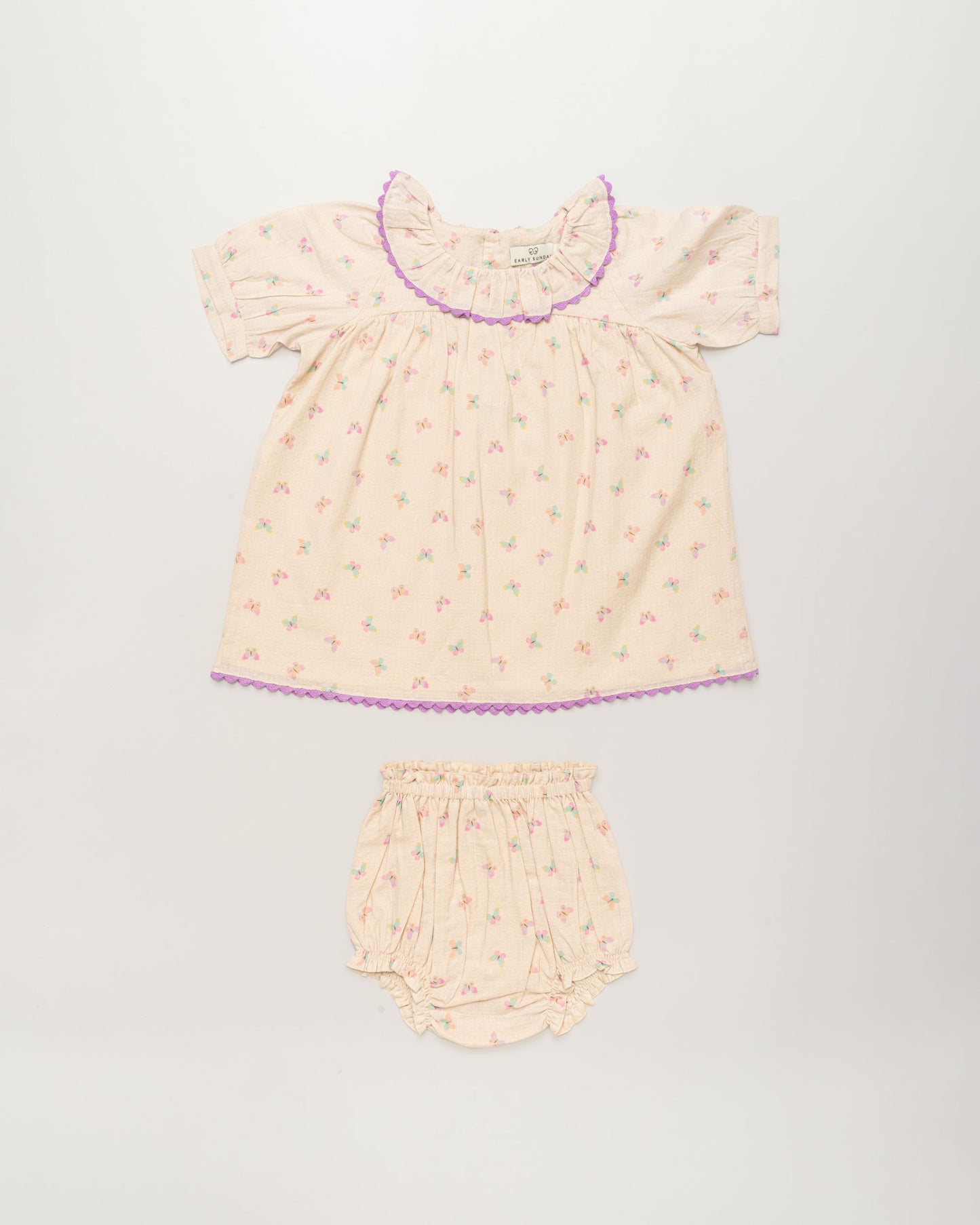 Early Sunday, Baby Bella Dress with Bloomer in Butterflies print, made from 100% Cotton Swiss Dobby Tiny Dots fabric, featuring short puff sleeves with a lilac lace & matching bloomers, shown in a front flat-lay view