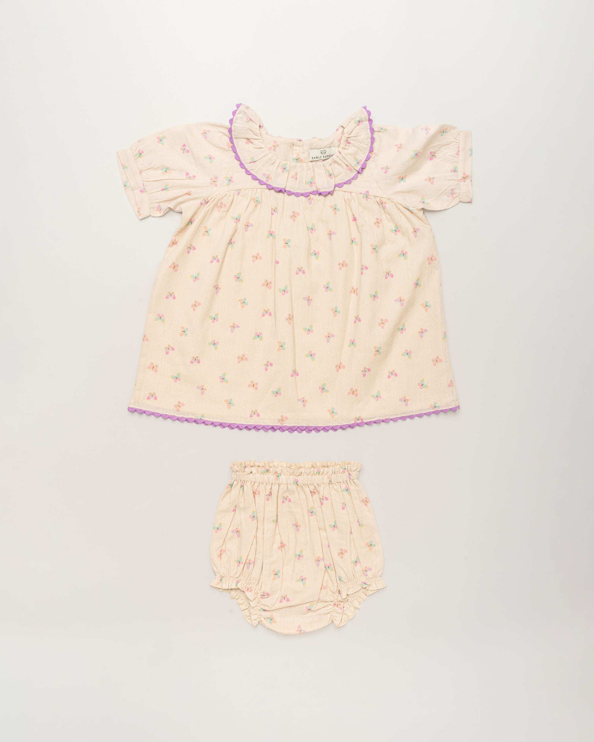 Early Sunday, Baby Bella Dress with Bloomer in Butterflies print, made from 100% Cotton Swiss Dobby Tiny Dots fabric, featuring short puff sleeves with a lilac lace & matching bloomers, shown in a front flat-lay view