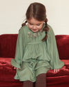 Early Sunday, Belle Dress in Sage Green, made from Viscose Twill fabric, featuring a square yoke bodice with puff sleeves and delicate gathered detailing, shown in a front view of model sitting on a sofa