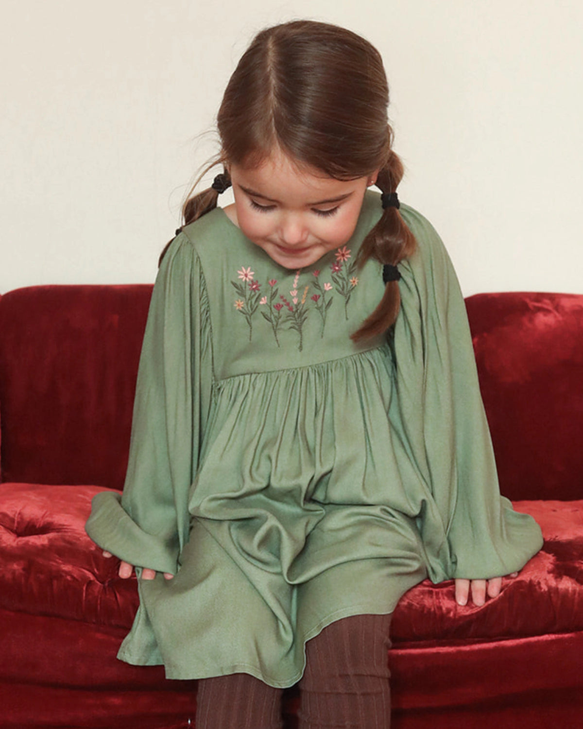 Early Sunday, Belle Dress in Sage Green, made from Viscose Twill fabric, featuring a square yoke bodice with puff sleeves and delicate gathered detailing, shown in a front view of model sitting on a sofa